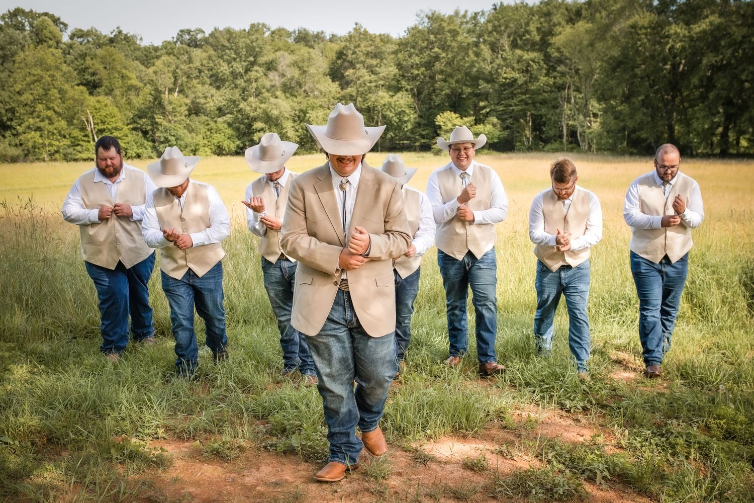 Groom in cowboy hat leads groomsmen in jeans and tan vests praying outdoors in a green field.