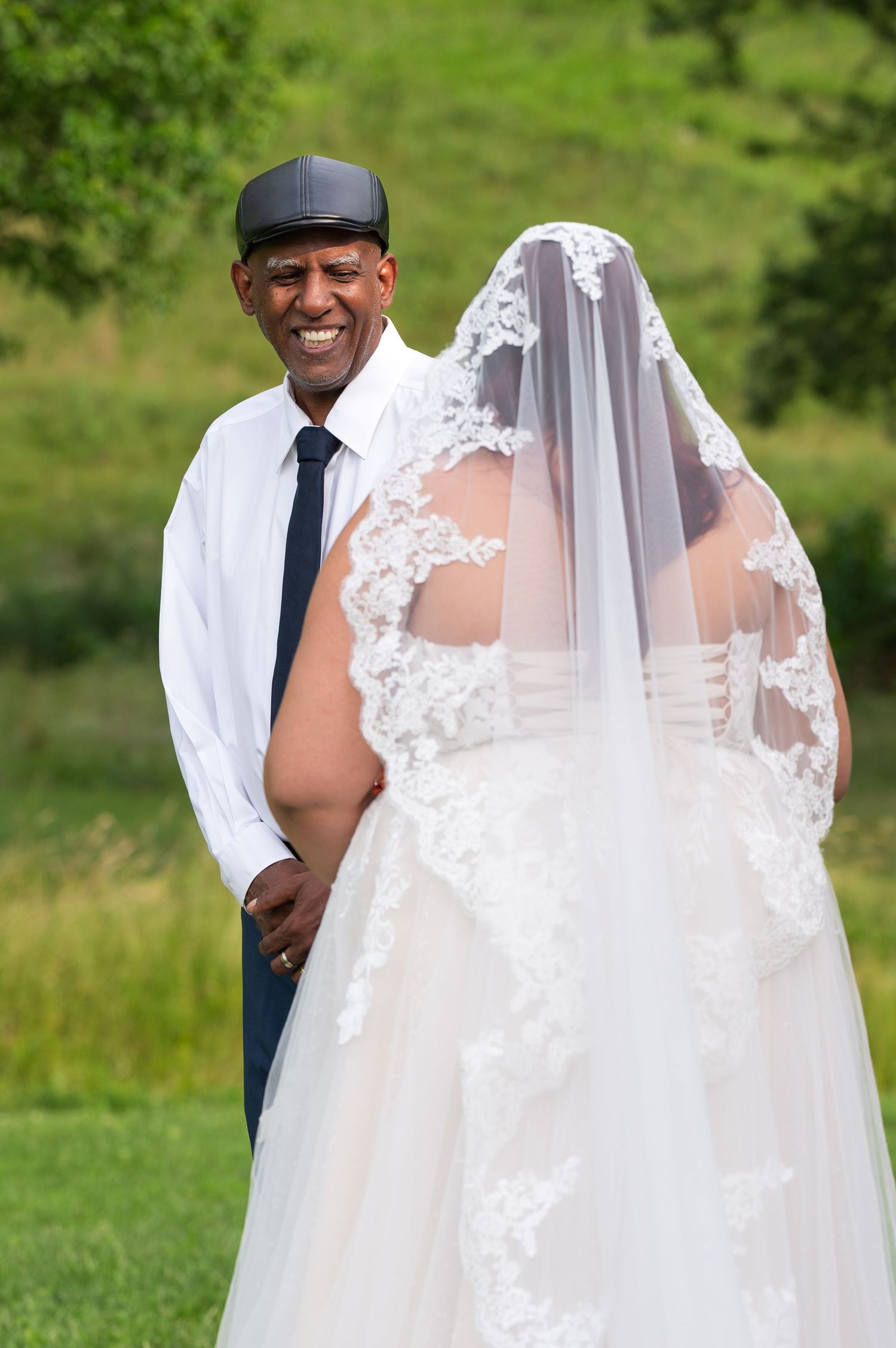Smiling groom in white shirt and cap stands beside bride in lace wedding gown and veil outdoors.