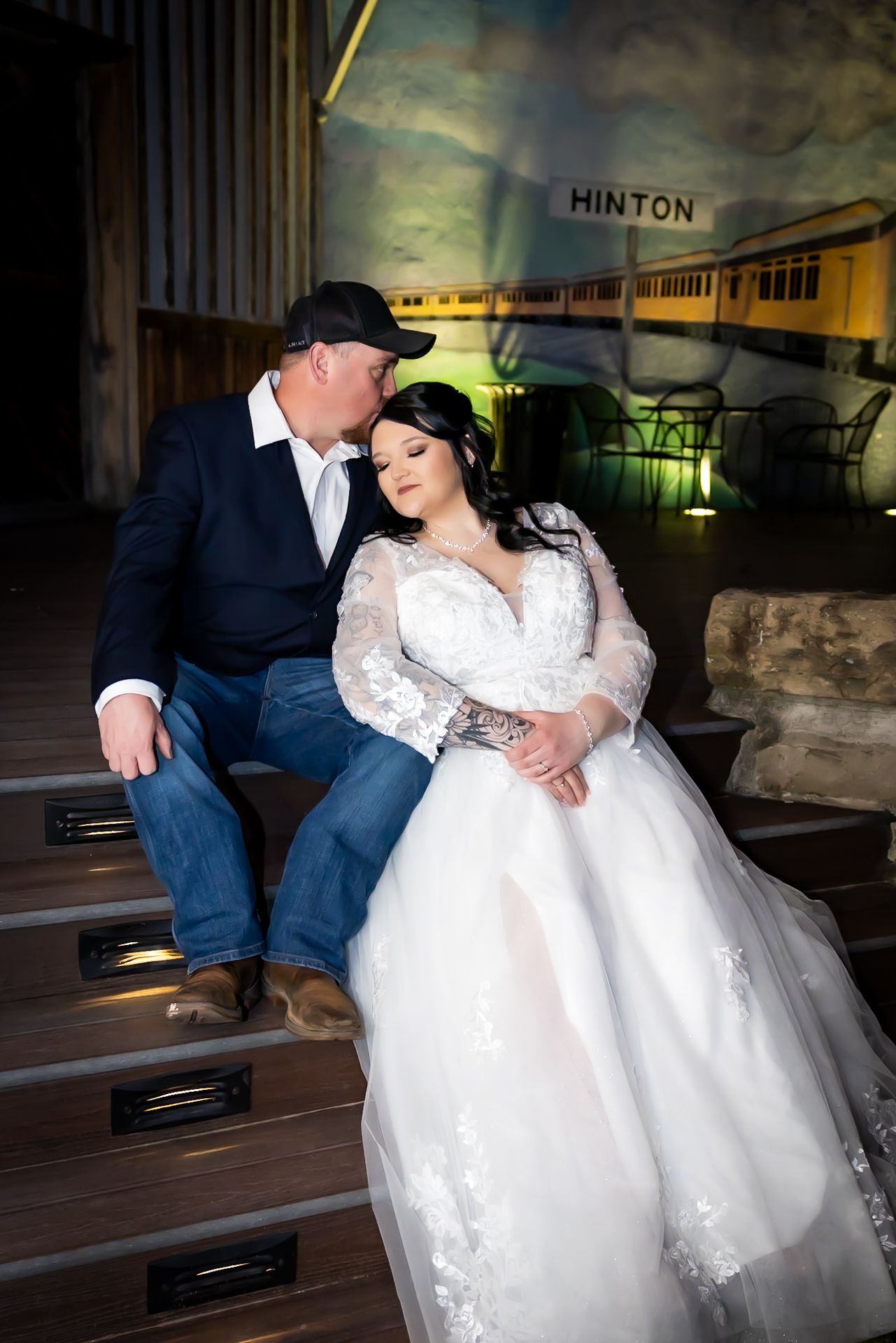 Bride in lace gown and groom in jeans sit together on steps at night.