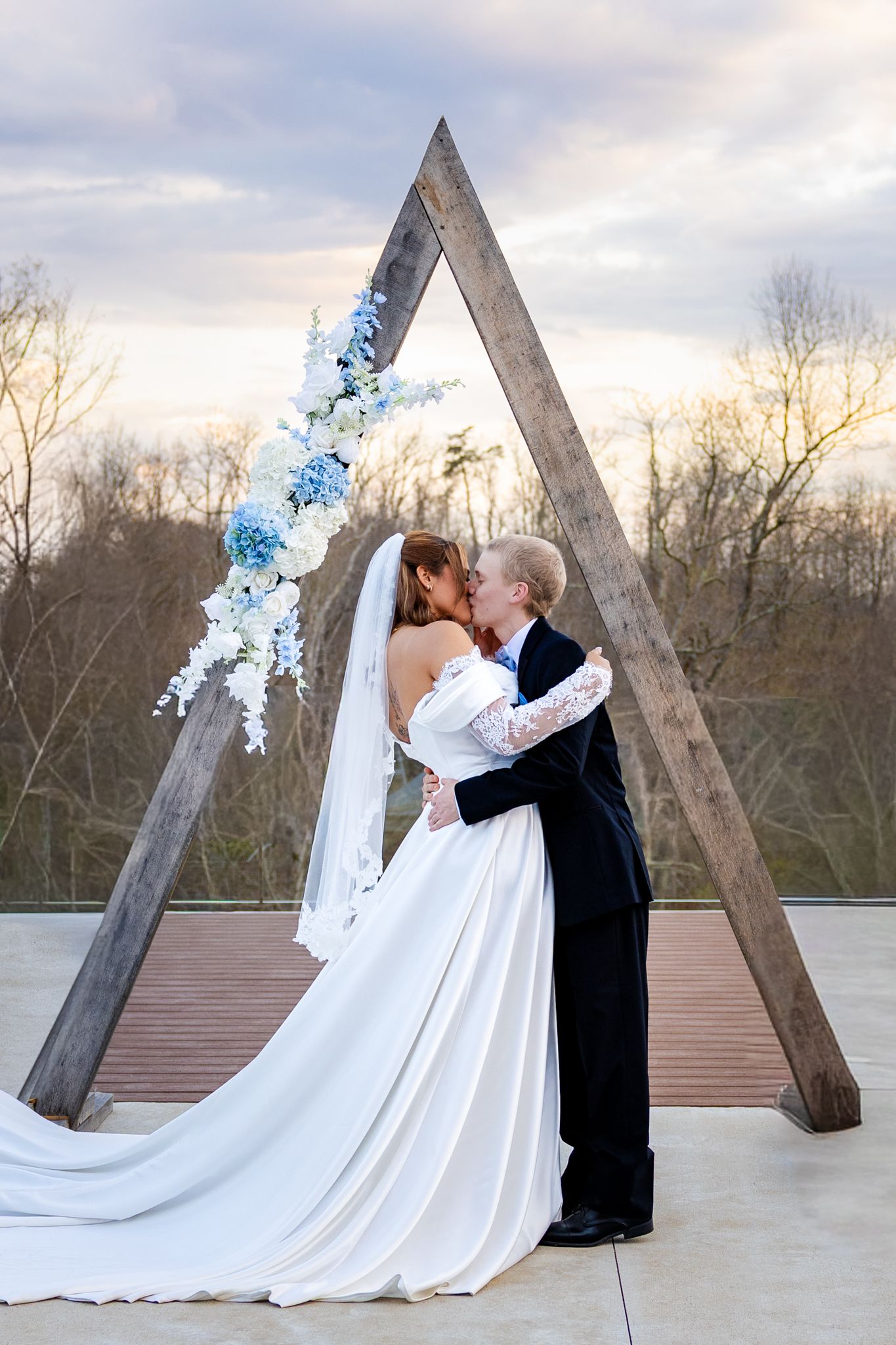 Newlyweds share first kiss under triangular wooden arch adorned with blue and white flowers.