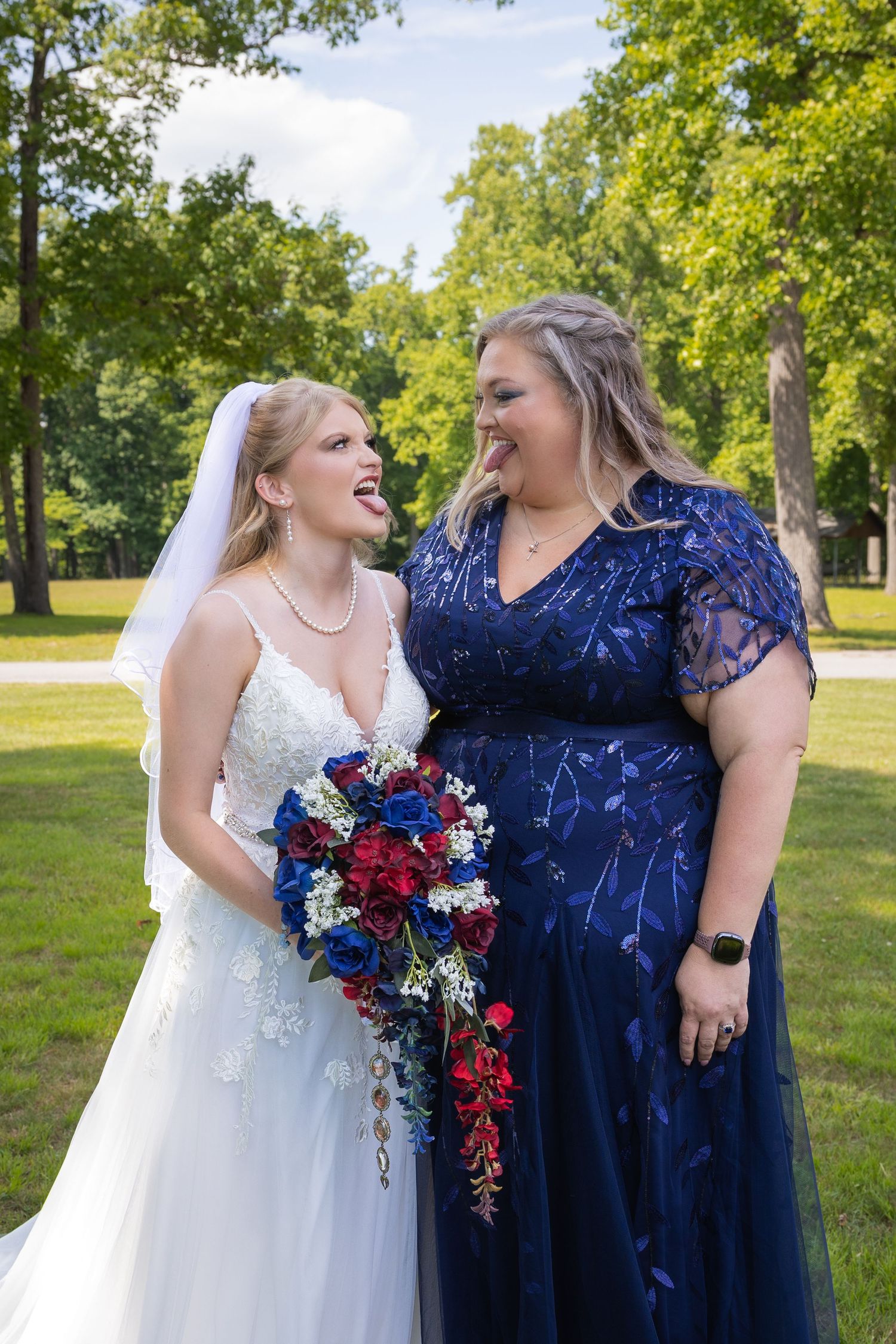 Bride in white gown with red and blue bouquet laughs with bridesmaid in navy dress outdoors.