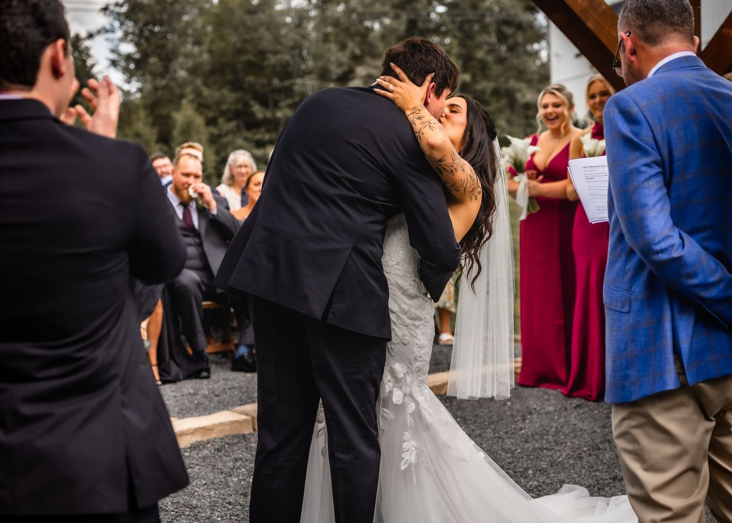 Bride and groom share their first kiss at outdoor wedding ceremony with guests and bridesmaid in red dress watching.