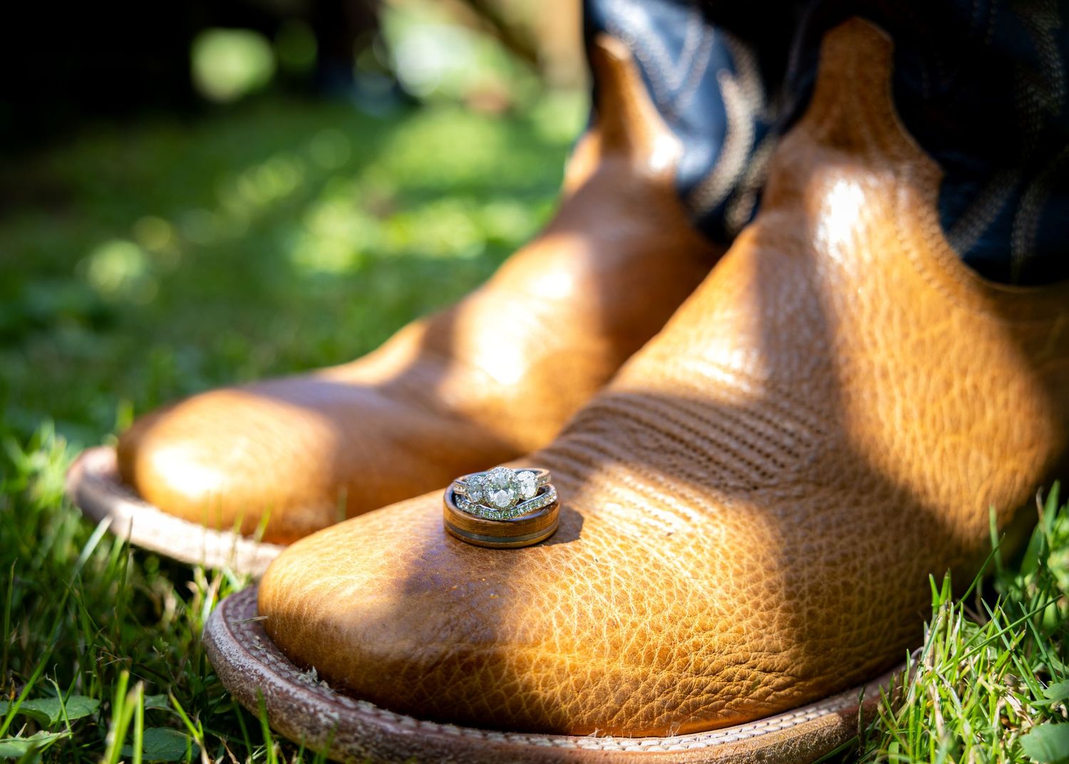 Close-up of tan cowboy boots on grass with wedding rings resting on the toe, perfect for a rustic wedding.