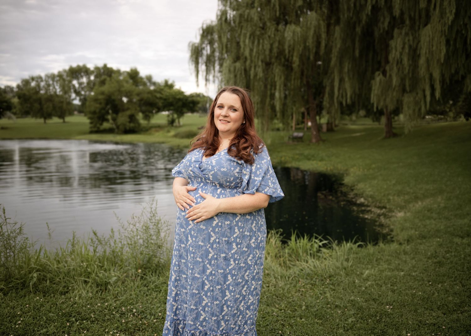 Pregnant woman in blue floral maxi dress cradling baby bump outdoors near a serene pond with weeping willow trees.