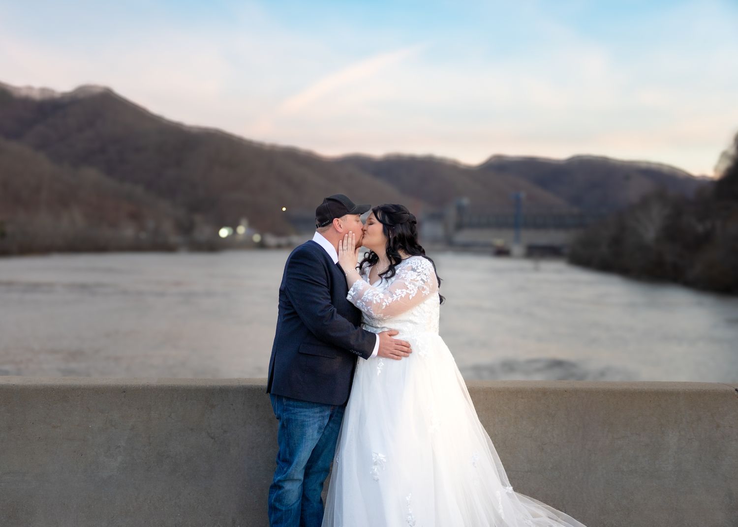Bride and groom share a tender kiss on a bridge with a scenic river and mountain backdrop at dusk.