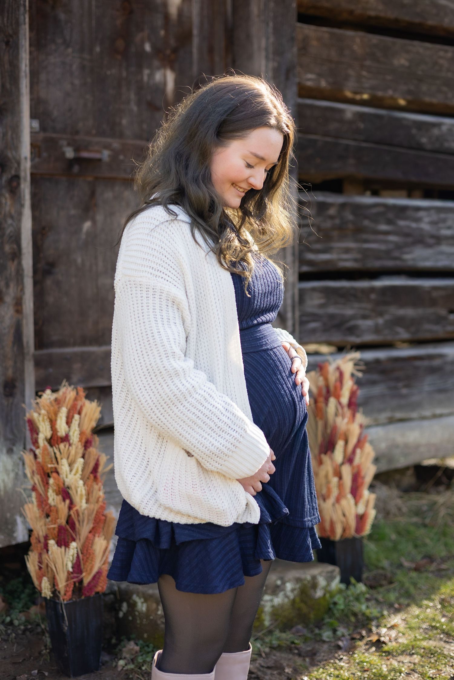 A pregnant woman in a white cardigan and blue dress cradles her baby bump beside a rustic wooden barn.