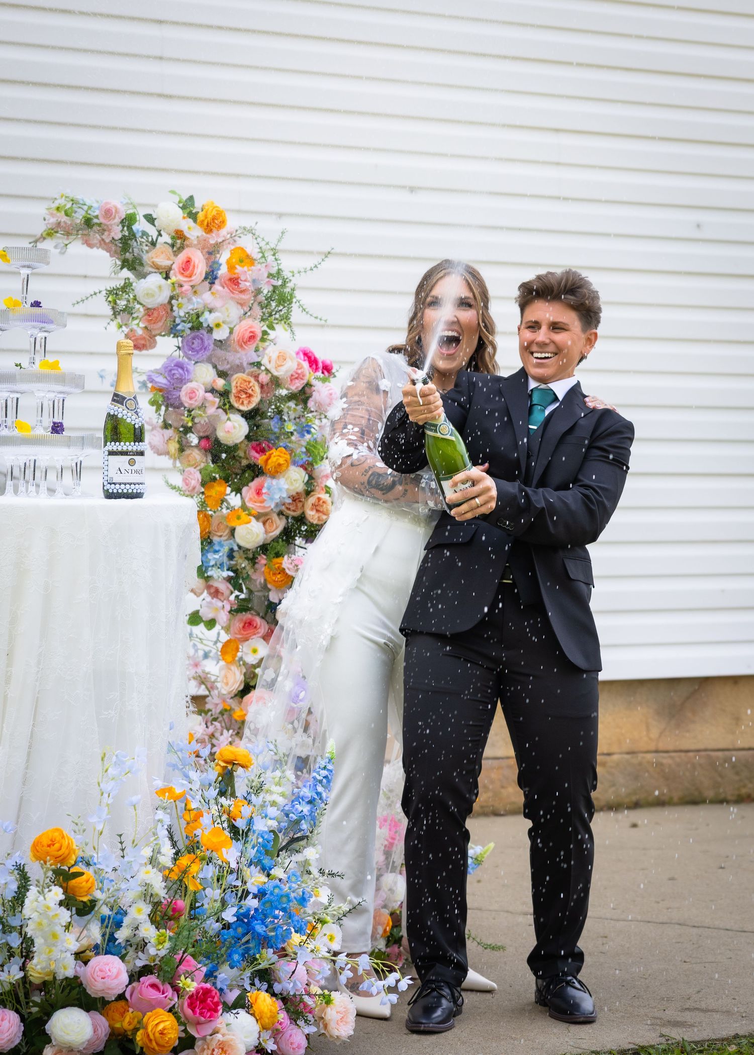 Joyful newlyweds popping champagne bottle beside colorful floral arch at wedding.