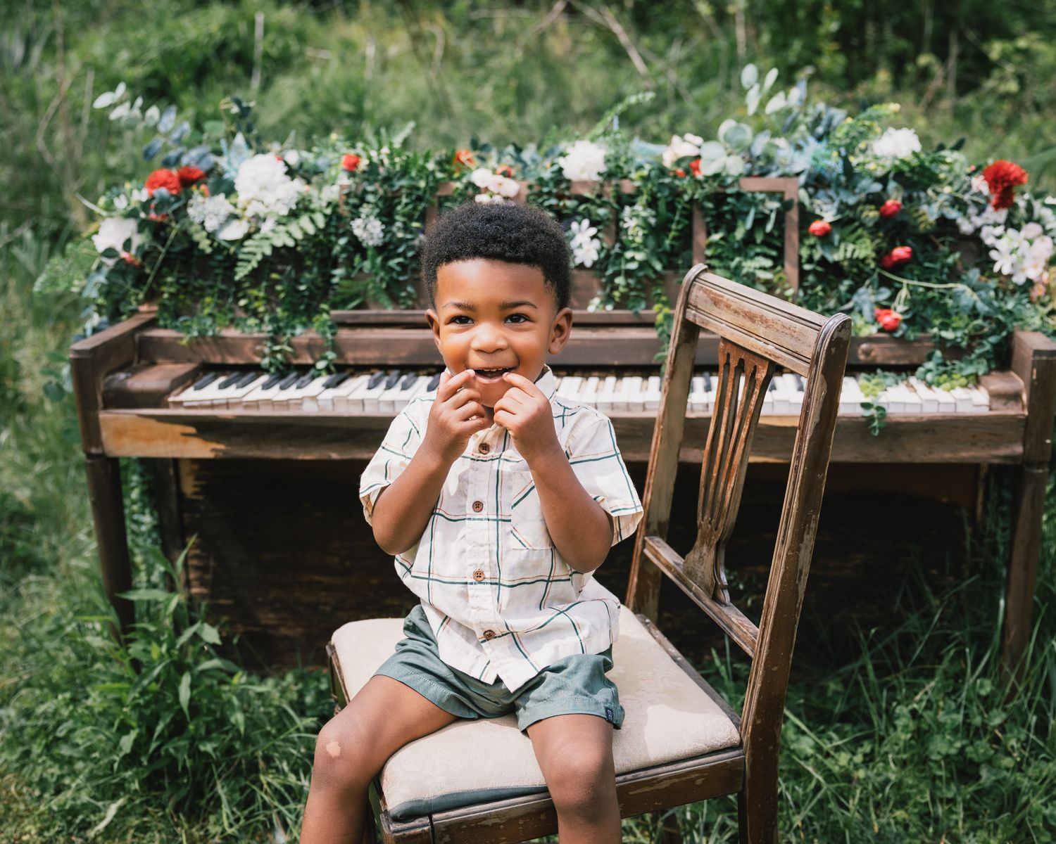 Happy young Black boy laughing and sitting on wooden chair in front of flower-adorned vintage piano outdoors.