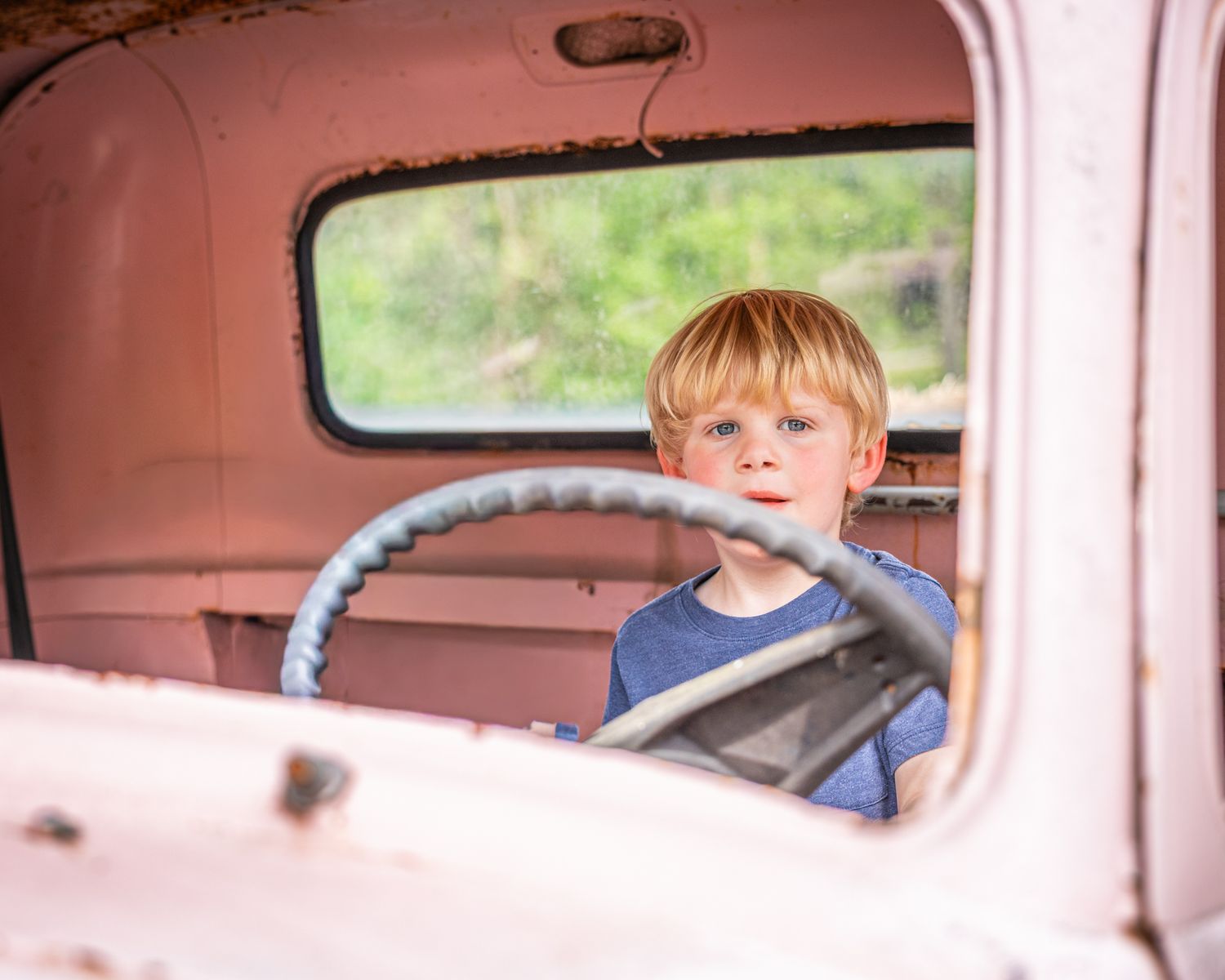 Curious red-haired boy sitting behind steering wheel of vintage rusty pink truck in summer.