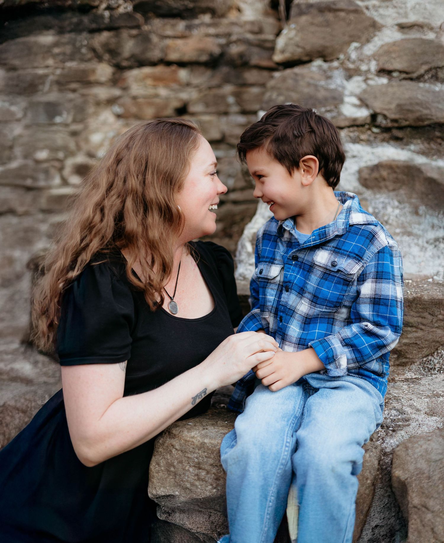 Mother in black dress smiling with young boy in blue plaid shirt on stone steps outdoors.