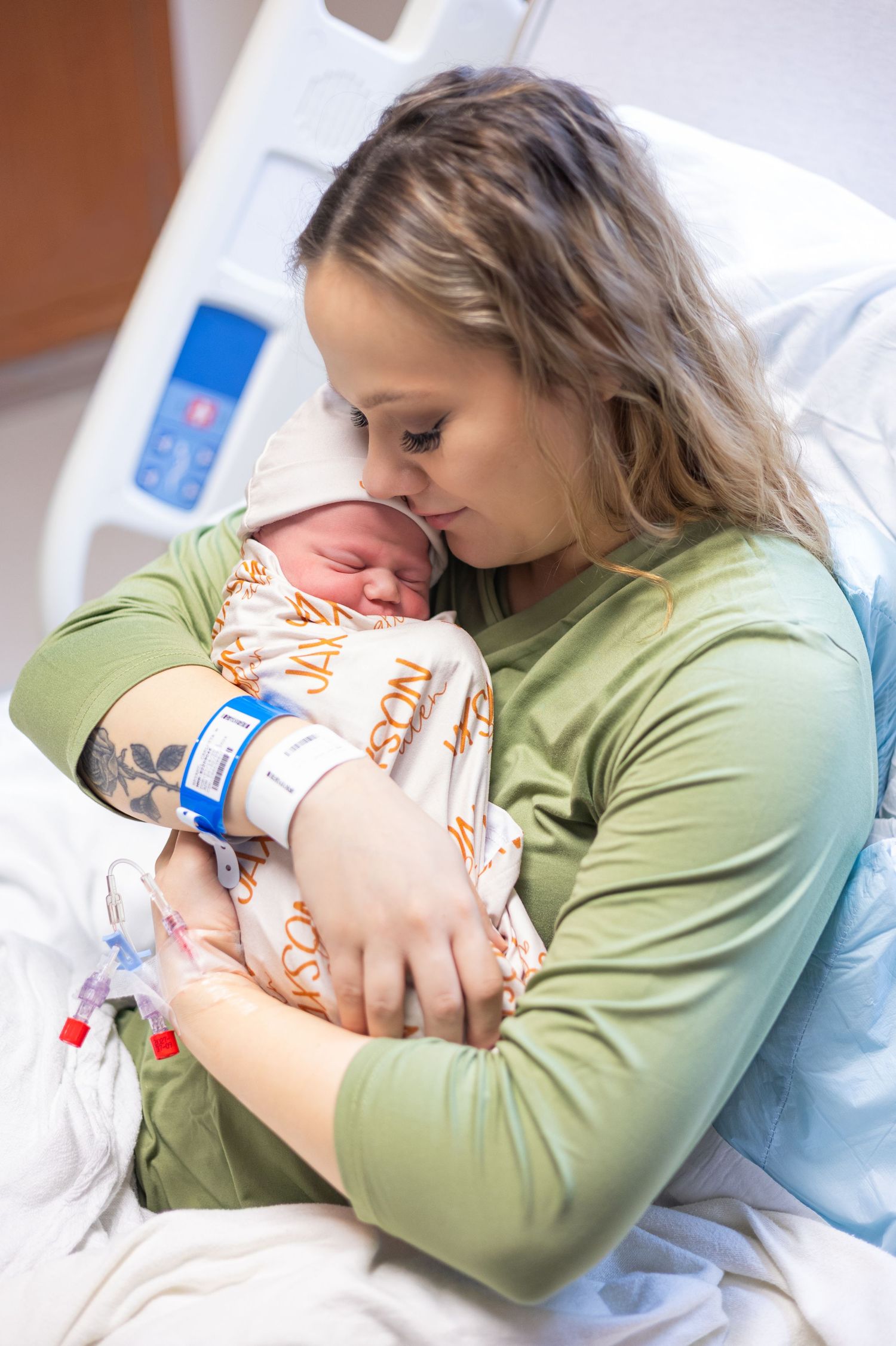 A tattooed mother in green shirt tenderly holds her swaddled newborn baby in hospital.