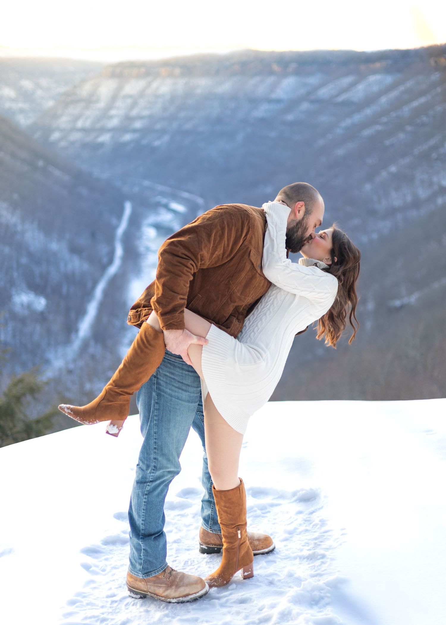 A couple shares a passionate kiss as the man dips the woman on a snowy mountain overlook at sunset.