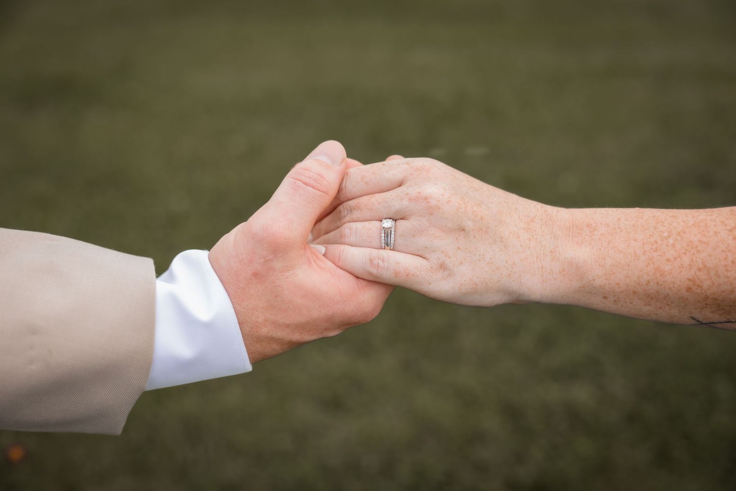 Close-up of a couple holding hands, woman wearing a diamond engagement ring, green grass background.