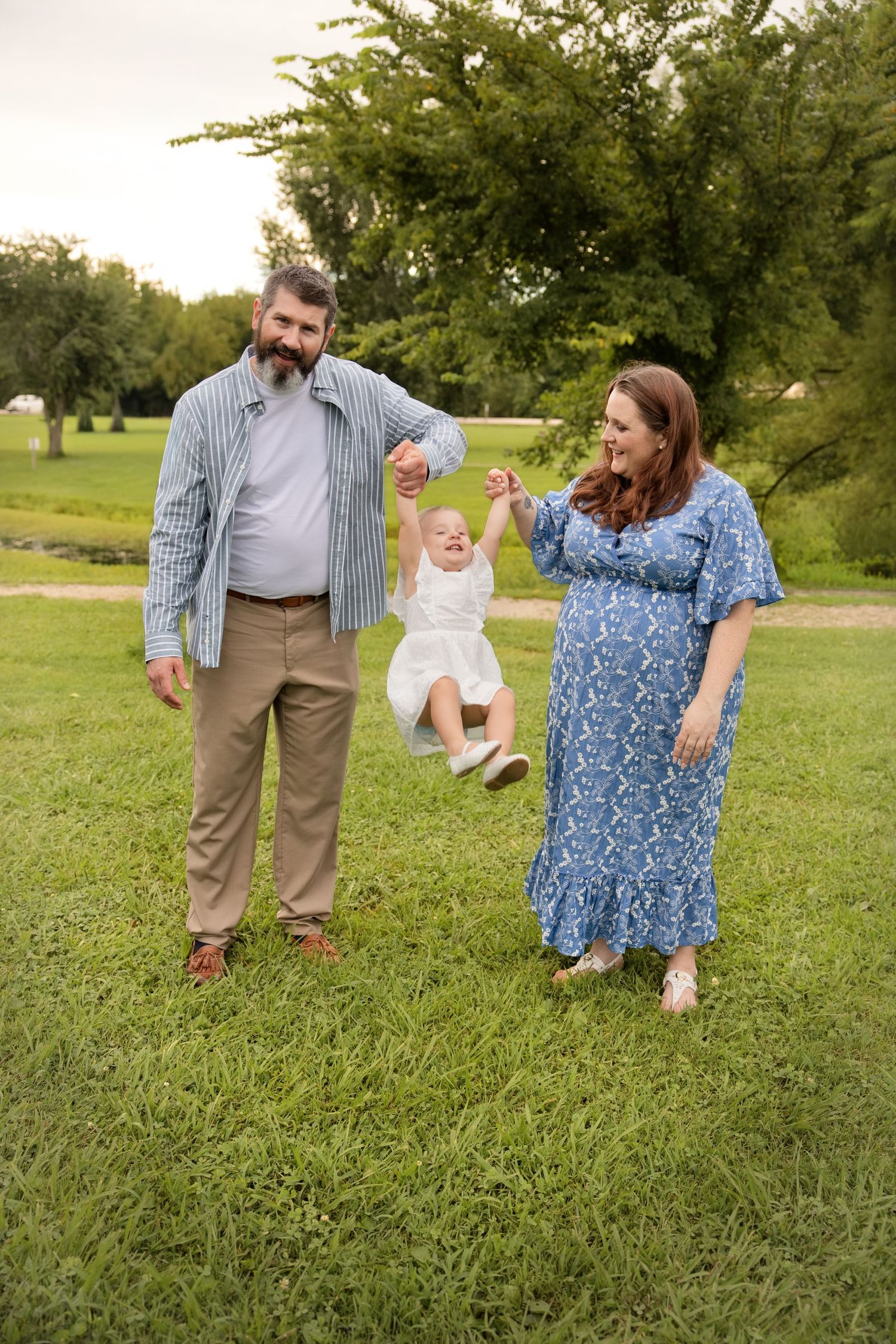 Happy family swinging toddler outdoors, pregnant mother in blue floral dress, bearded father in striped shirt, green park.