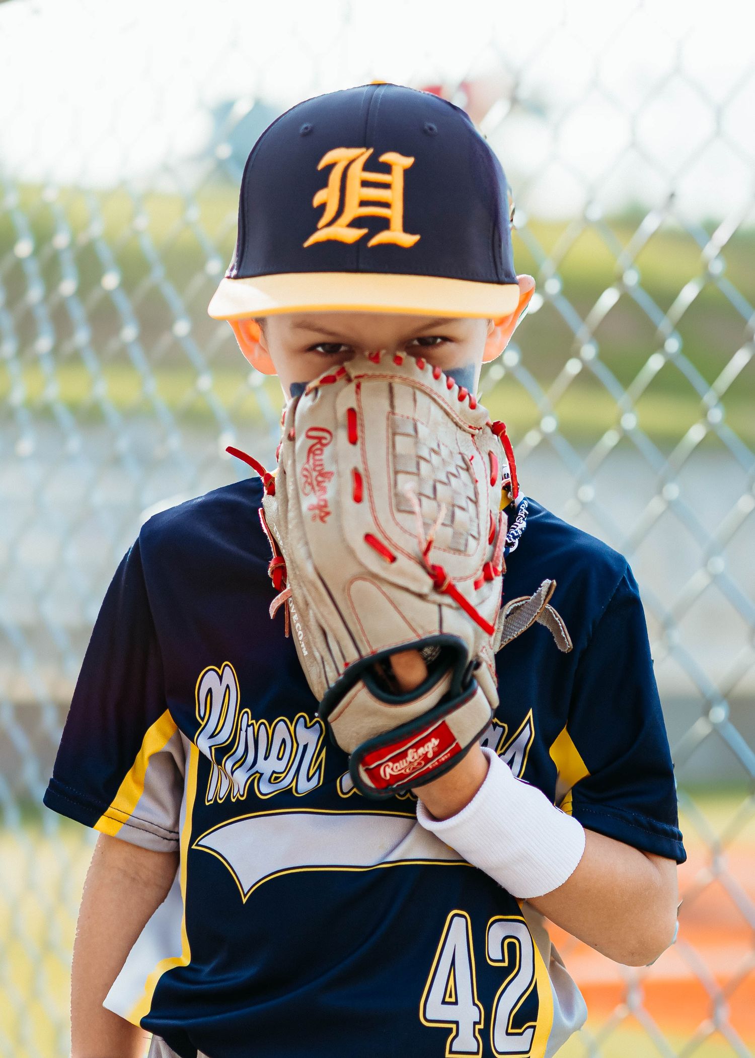 Young baseball player in navy uniform number 42 hiding face behind Rawlings glove on field.