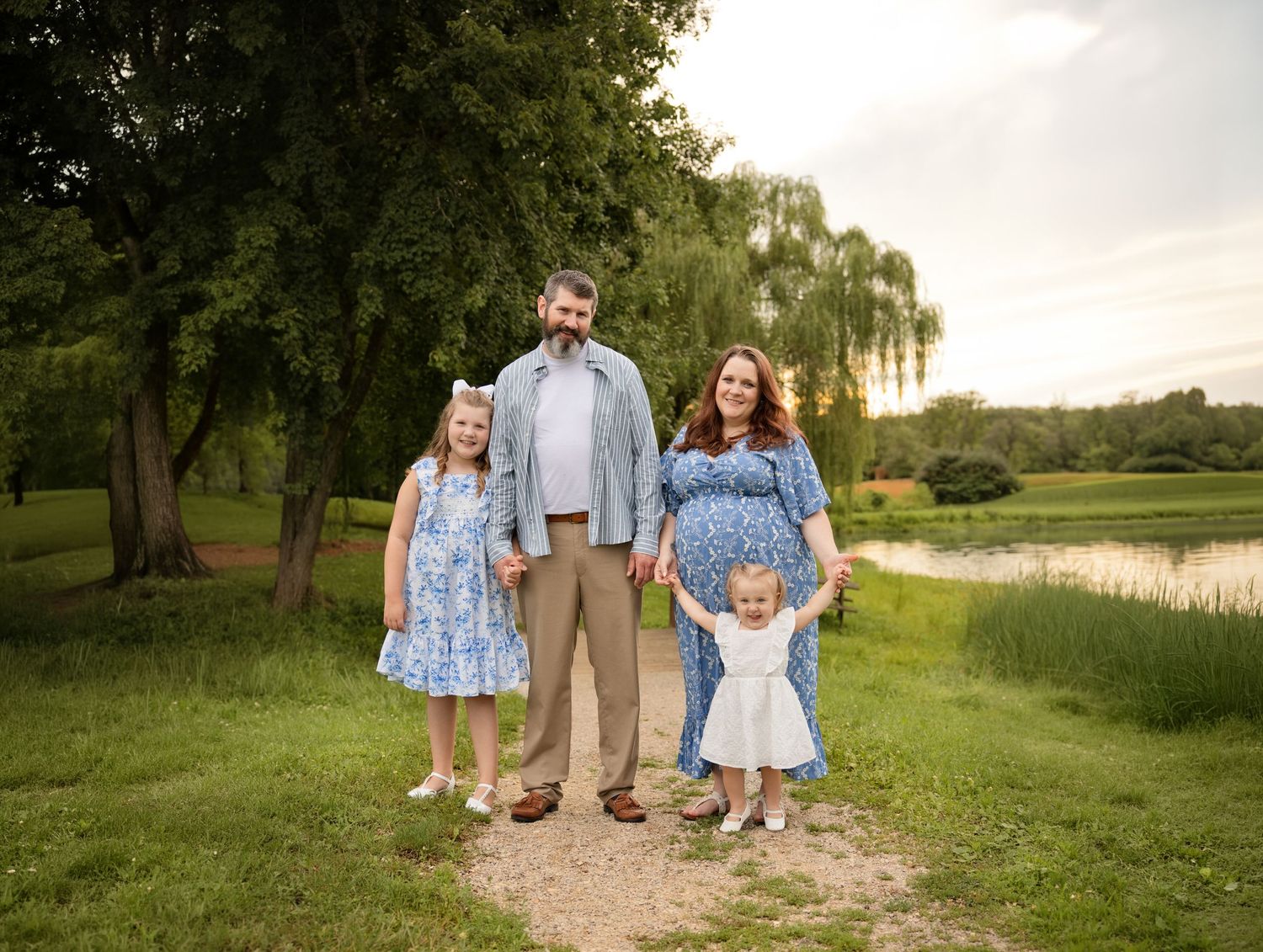 Family of four poses outdoors near a pond, with pregnant mother, father, and two young daughters in matching blue dresses.