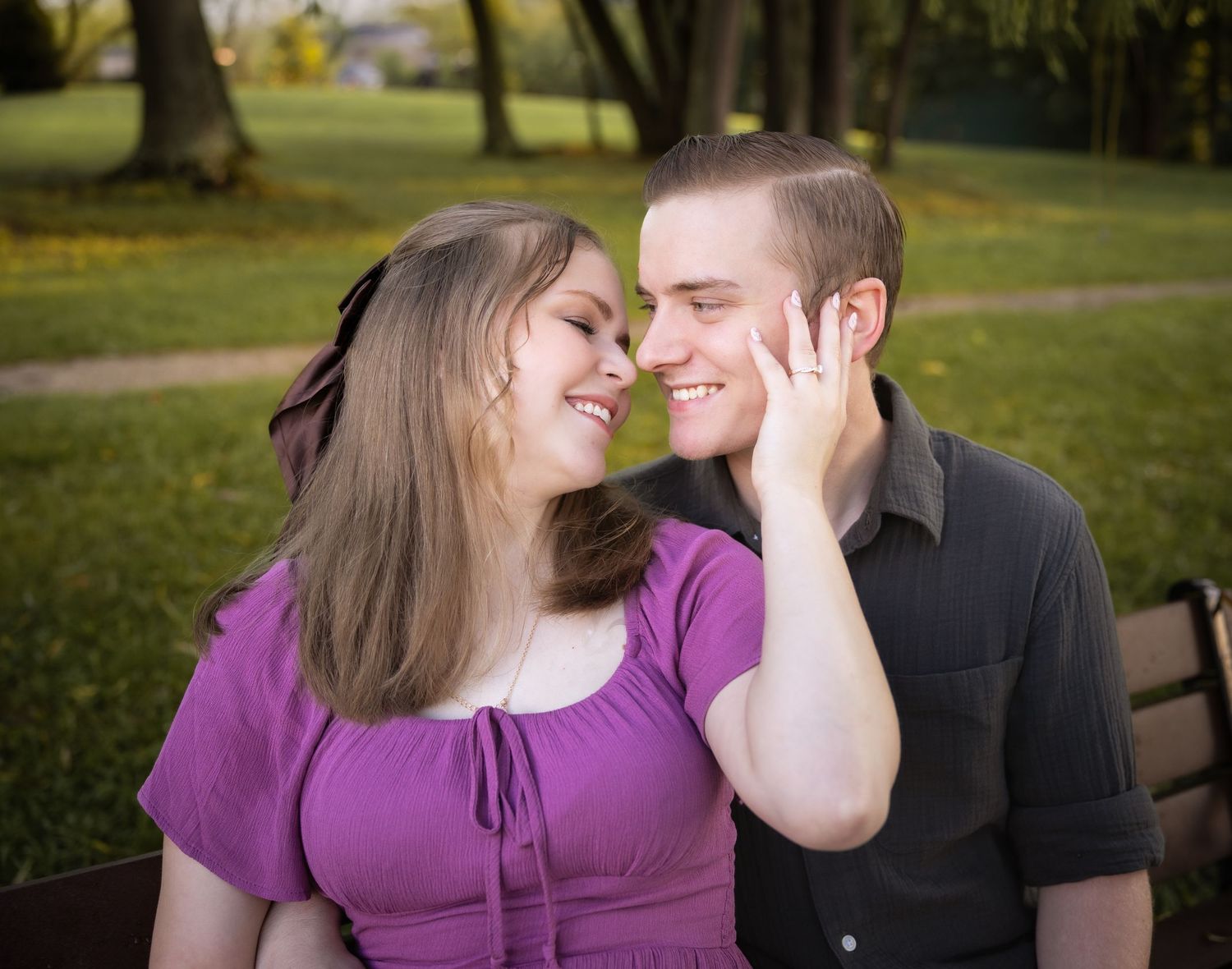 A young couple shares a romantic moment outdoors, smiling at each other in a lush green park setting.