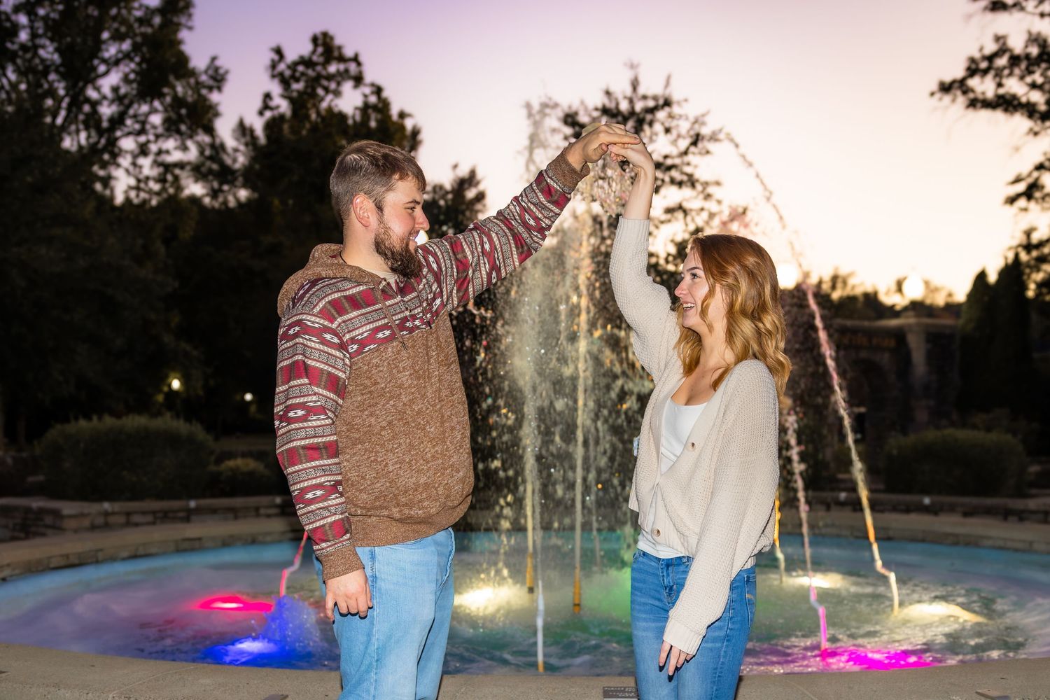 Smiling couple dances beside a glowing illuminated fountain at dusk as the man twirls the woman in a romantic evening engagement photo.
