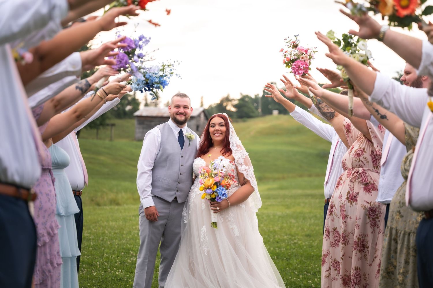 Bride and groom walk through floral bouquet arch held by wedding guests on green lawn.