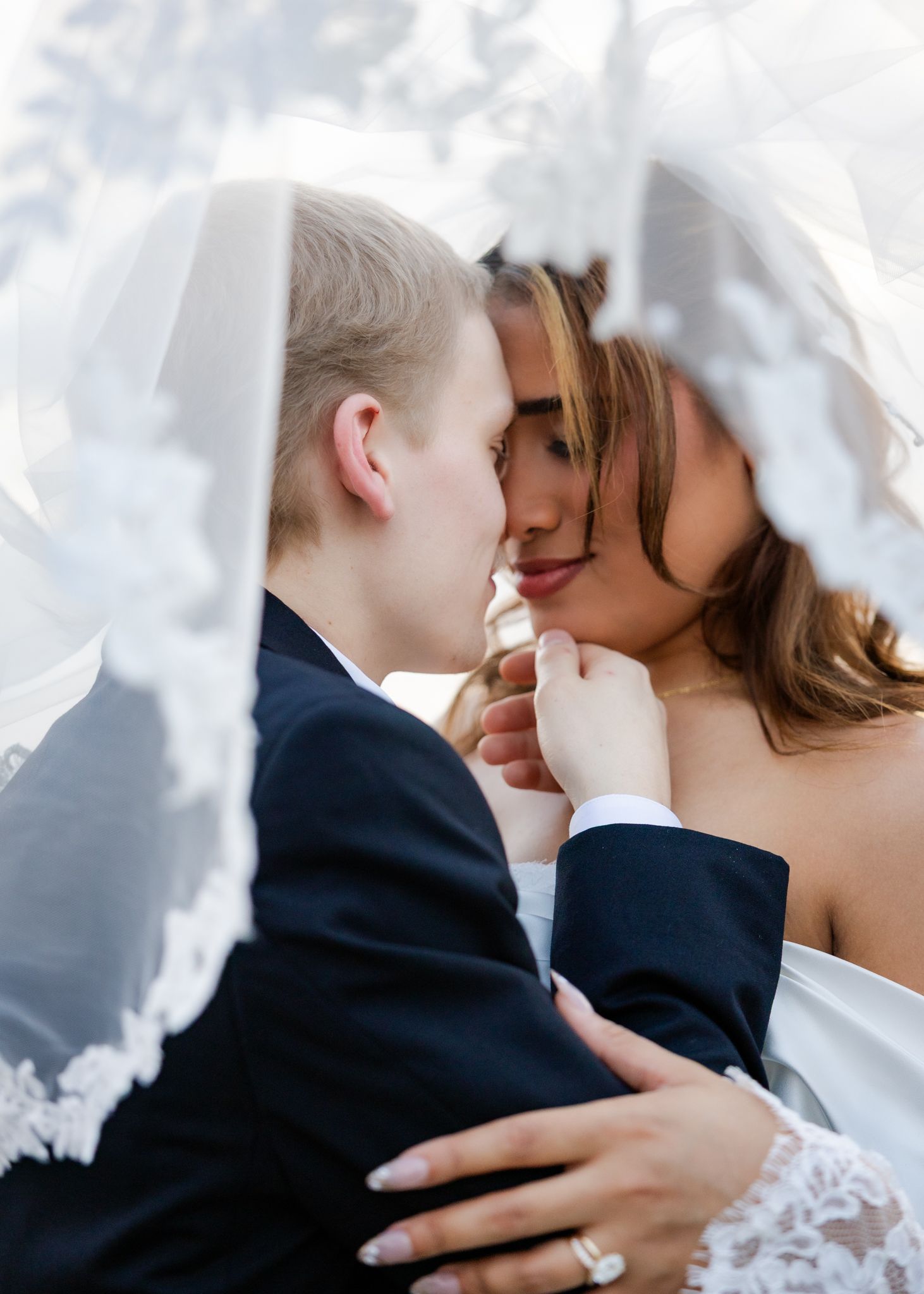 Couple shares intimate moment under lace bridal veil during outdoor wedding photo session.