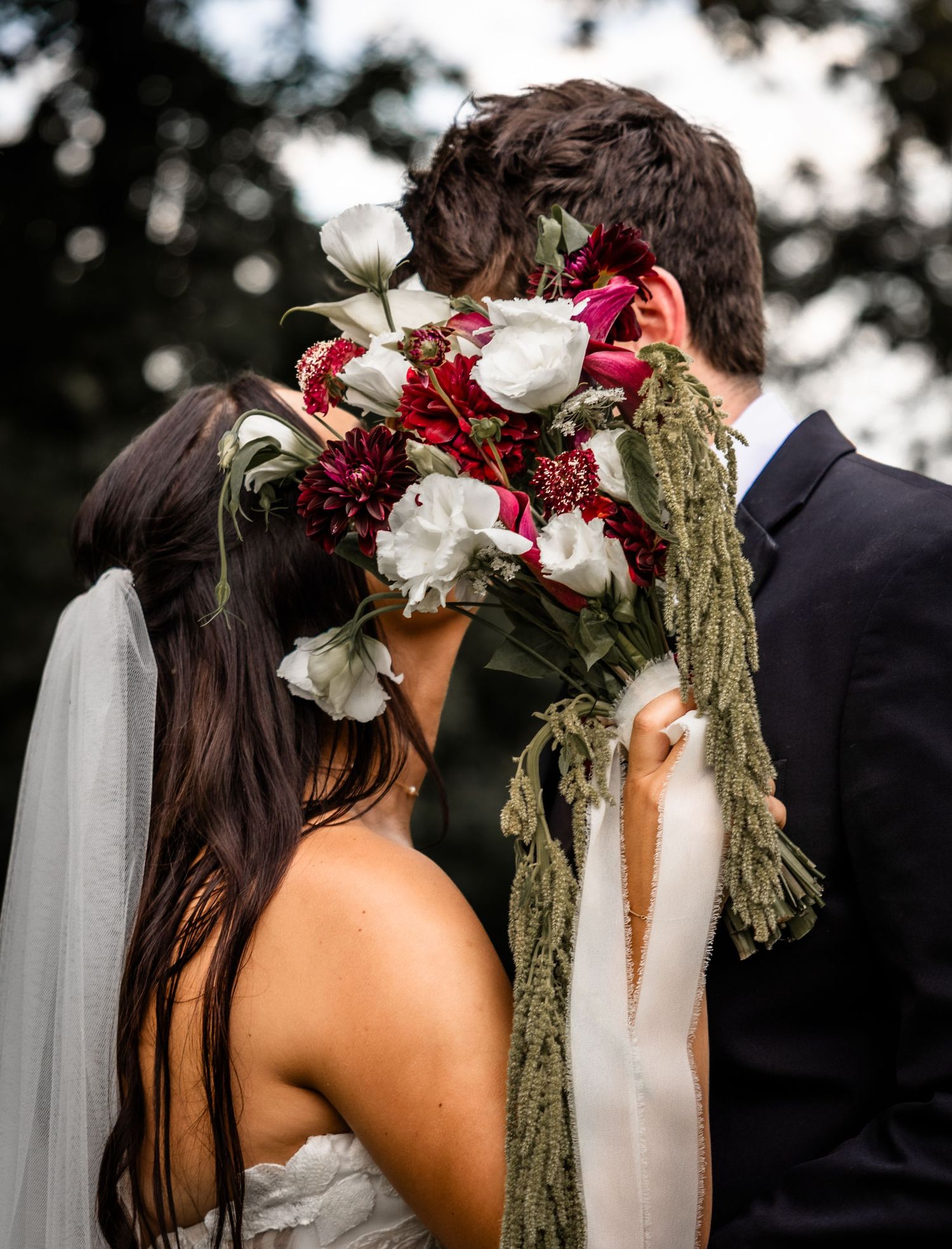 Bride and groom kissing behind a lush bridal bouquet of white and red flowers with green trailing accents.