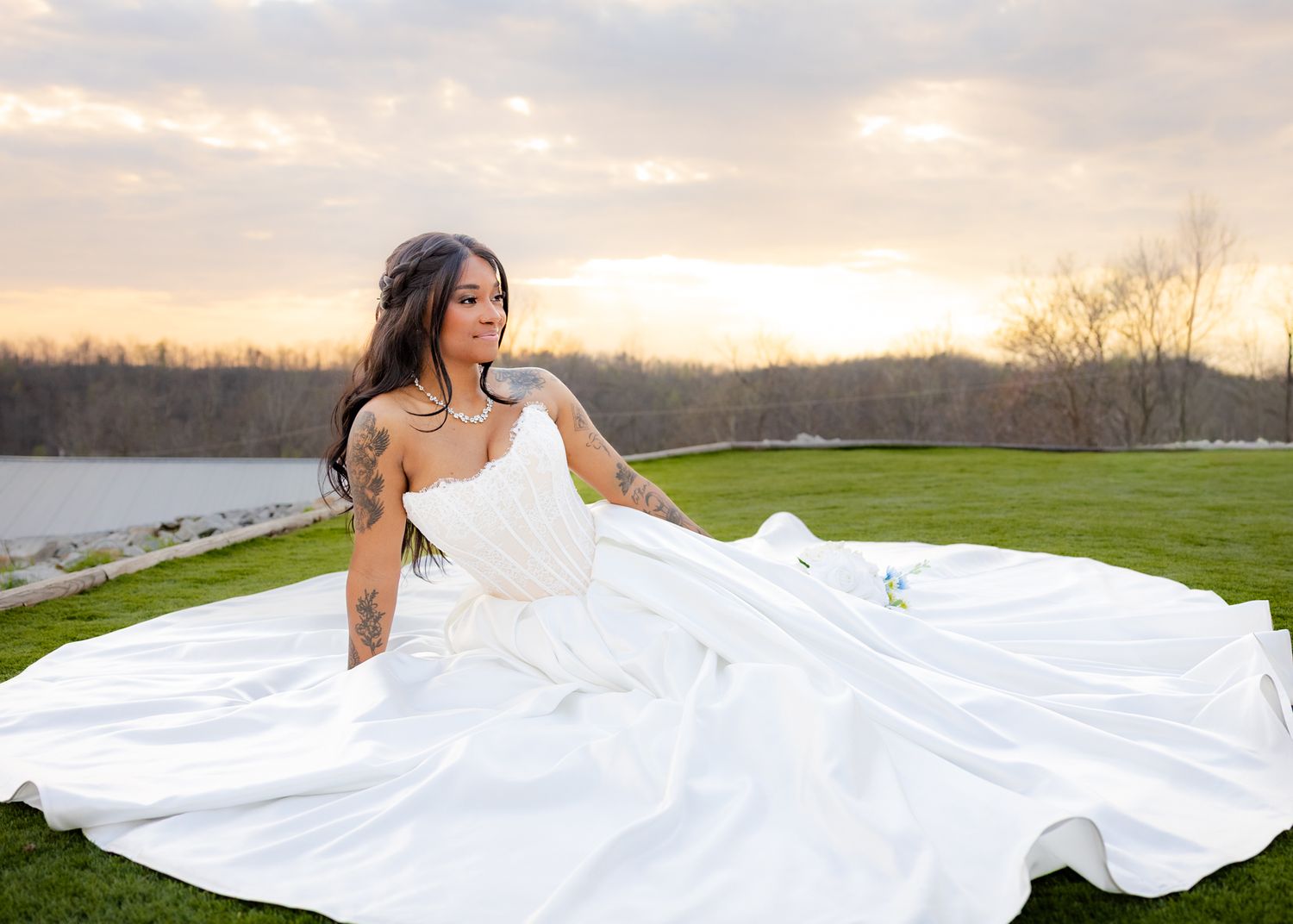 Tattooed bride in flowing white gown sits on lawn at sunset beside a serene lake.