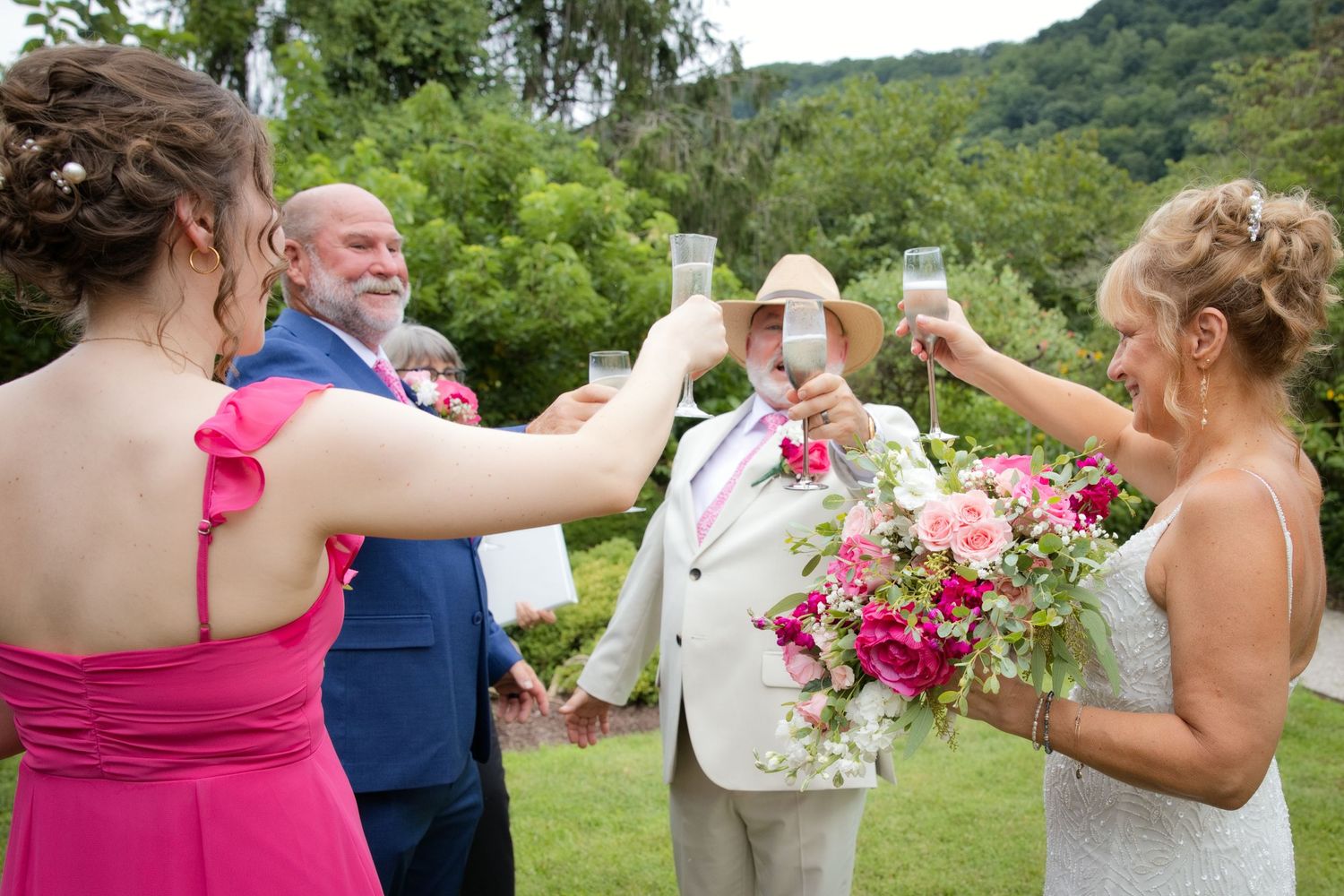 Wedding party toasting with champagne glasses outdoors, bride holding pink floral bouquet in elegant garden ceremony setting.