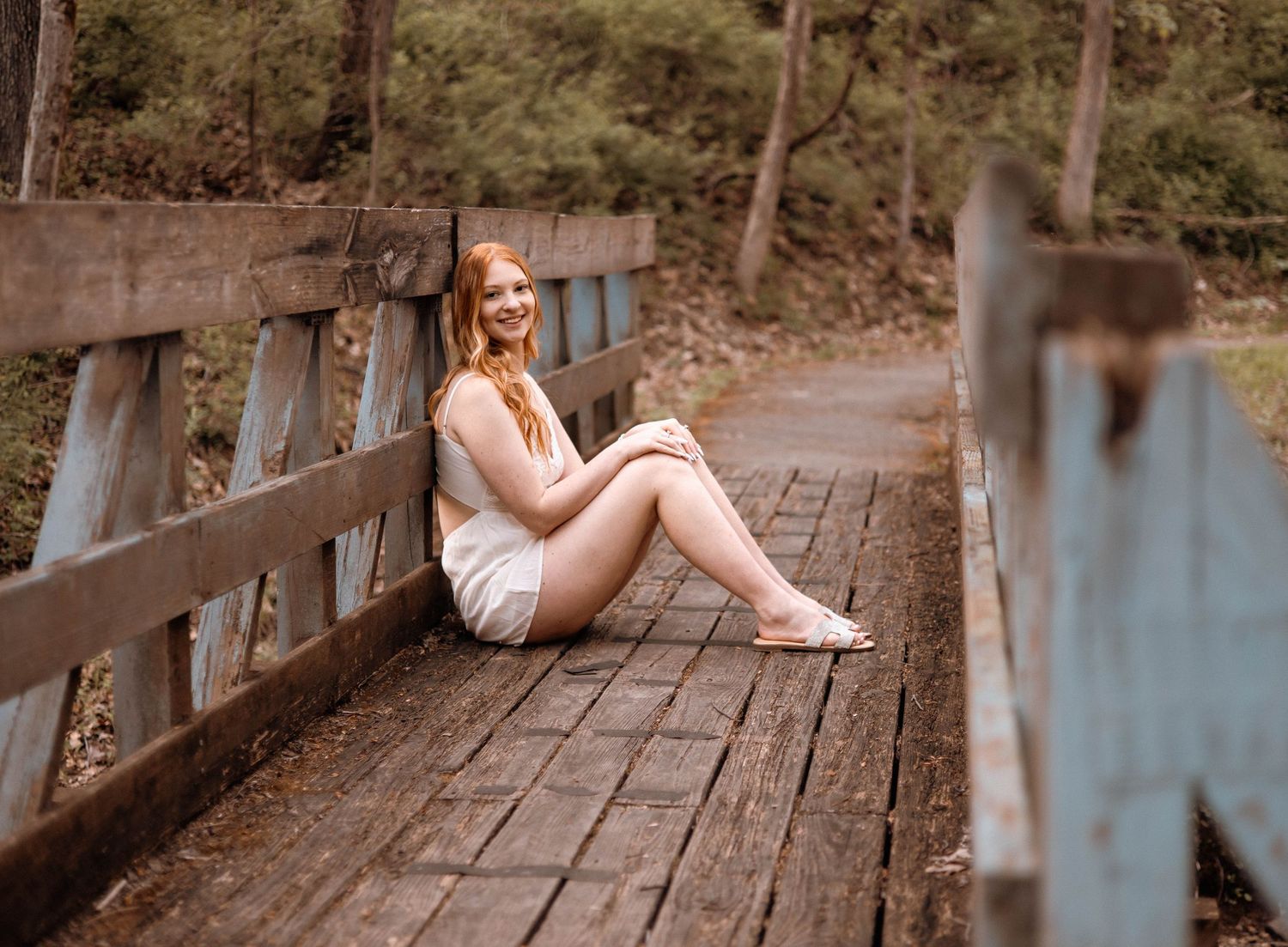 Smiling redhead woman in white shorts sitting on rustic wooden bridge in wooded outdoor setting.