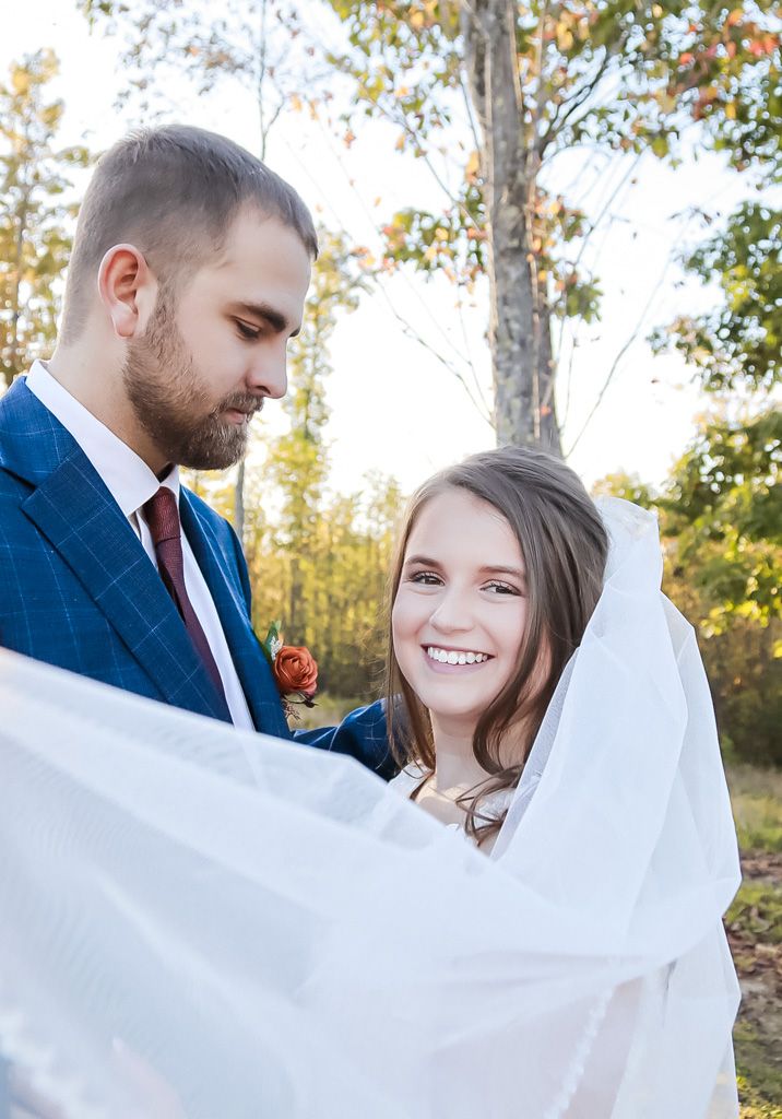 Groom wrapping bride in white veil as she smiles joyfully in autumn forest setting.