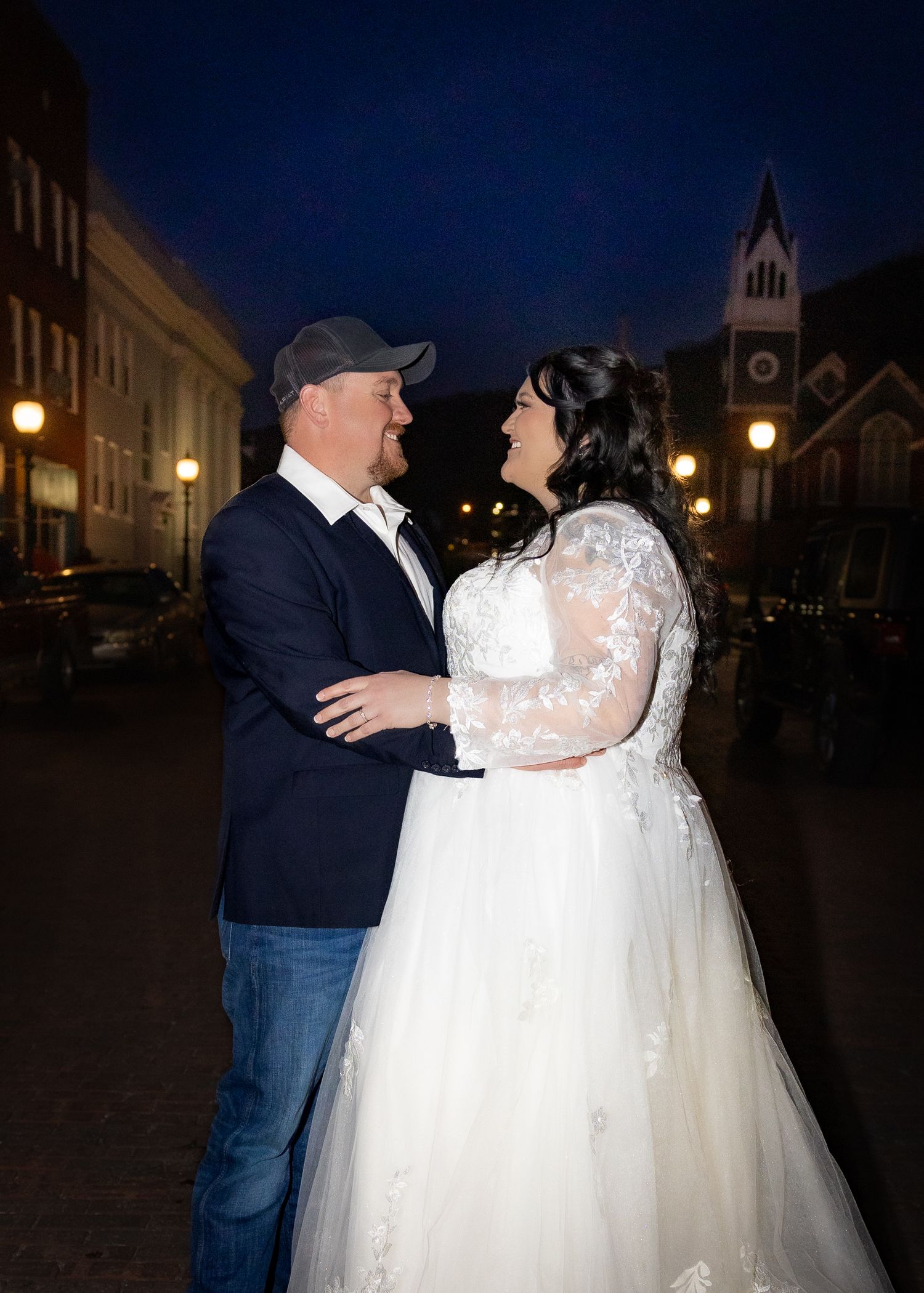 Bride and groom embrace and smile at each other on a lit city street at night.