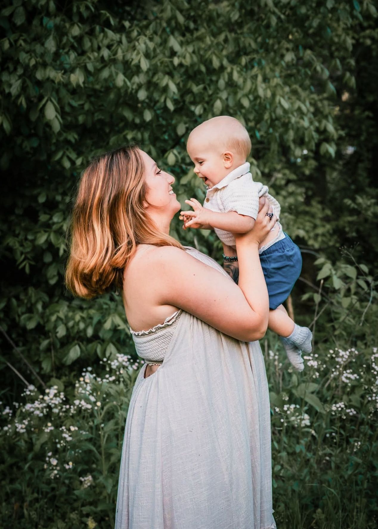 A mother in a flowy dress holds her smiling baby outdoors among wildflowers and green trees.