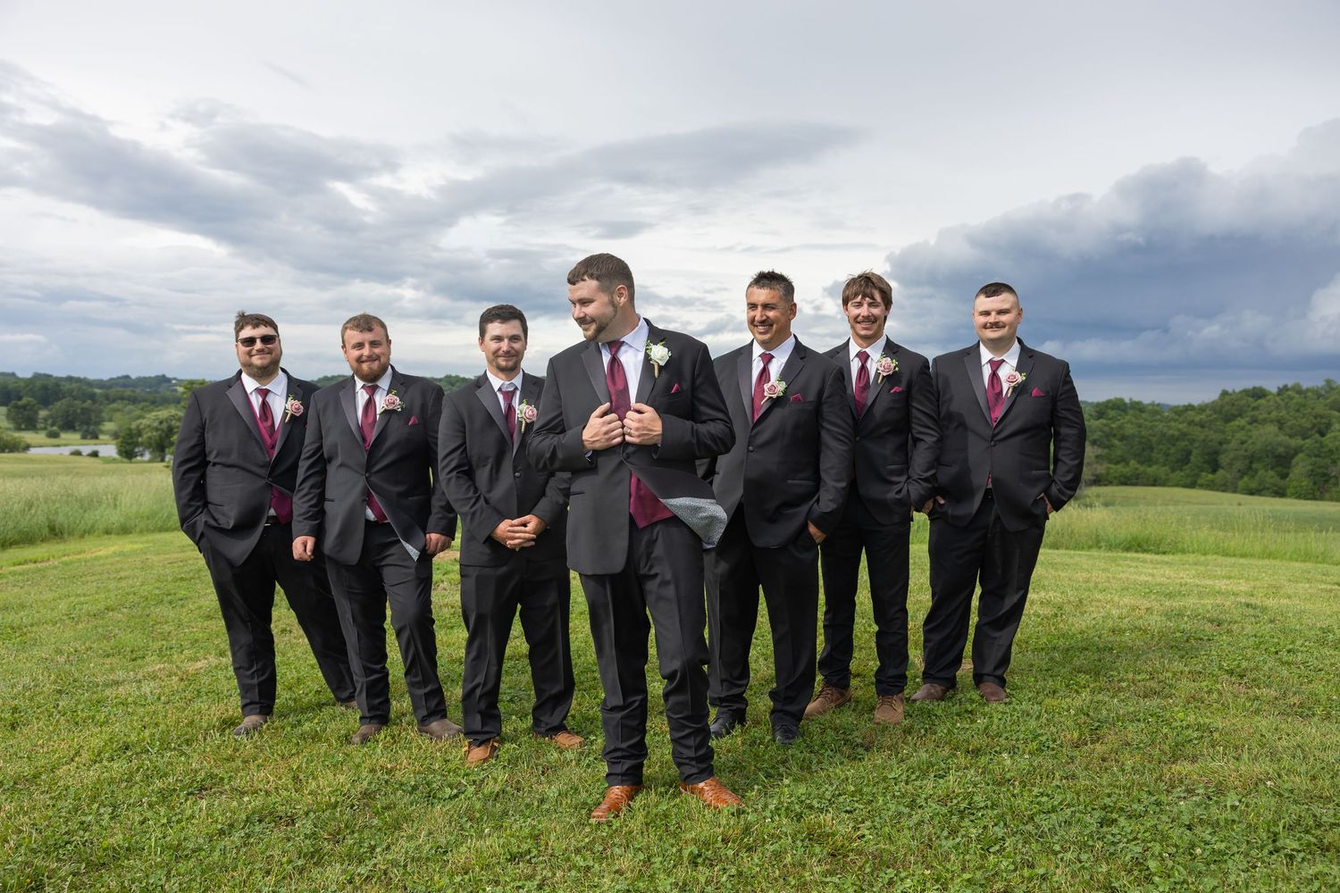 Groomsmen in dark suits with burgundy ties posing on a green hillside under a cloudy sky.