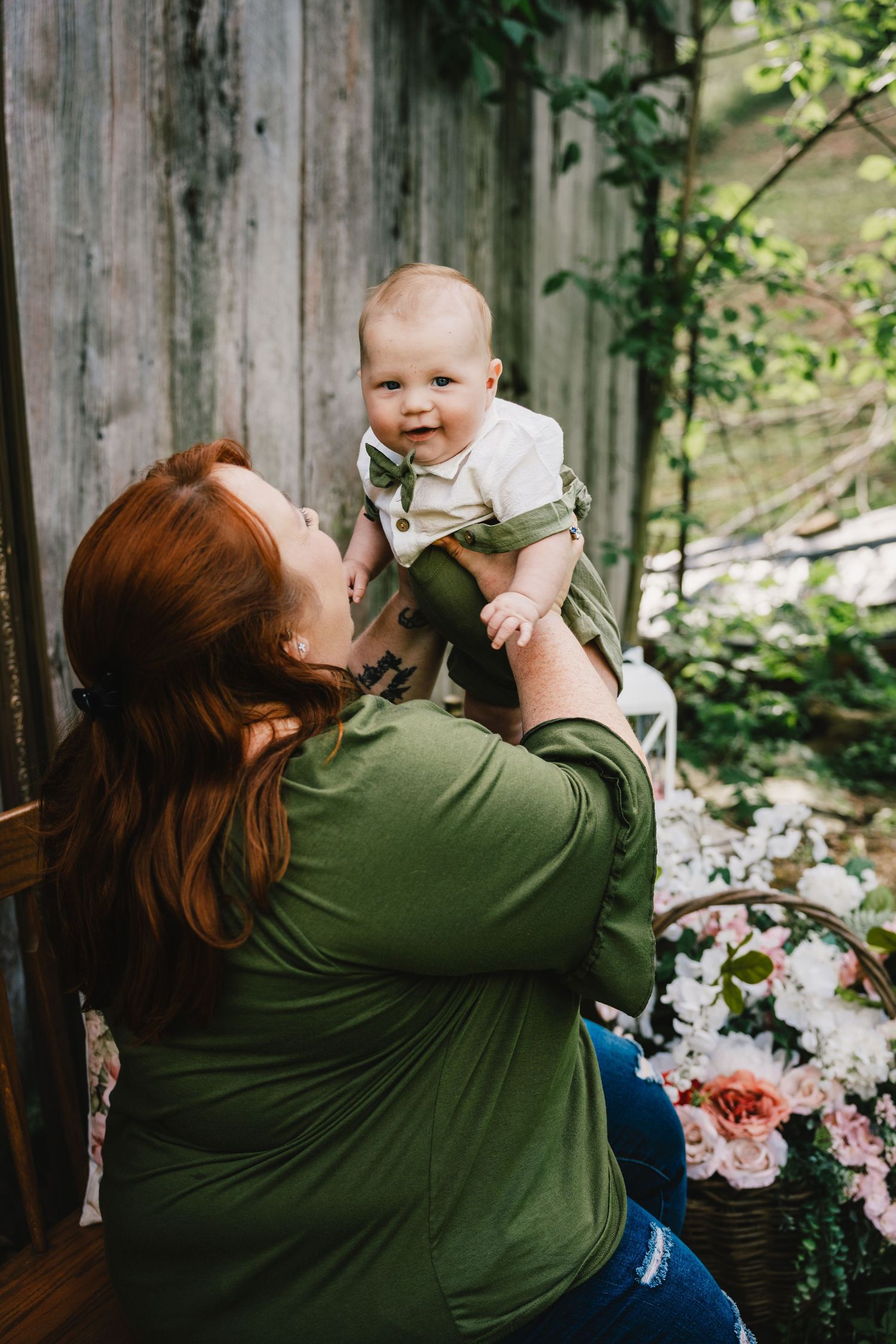 Red-haired woman in green top lifting smiling baby boy outdoors near rustic wood wall and flowers.