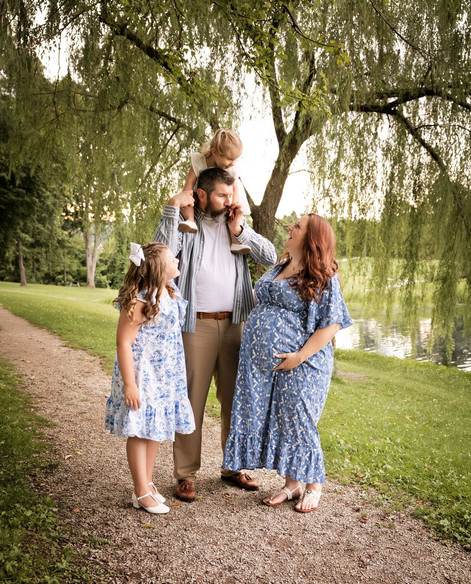 Pregnant woman in blue floral dress poses with husband and two young daughters by a pond under willow trees.