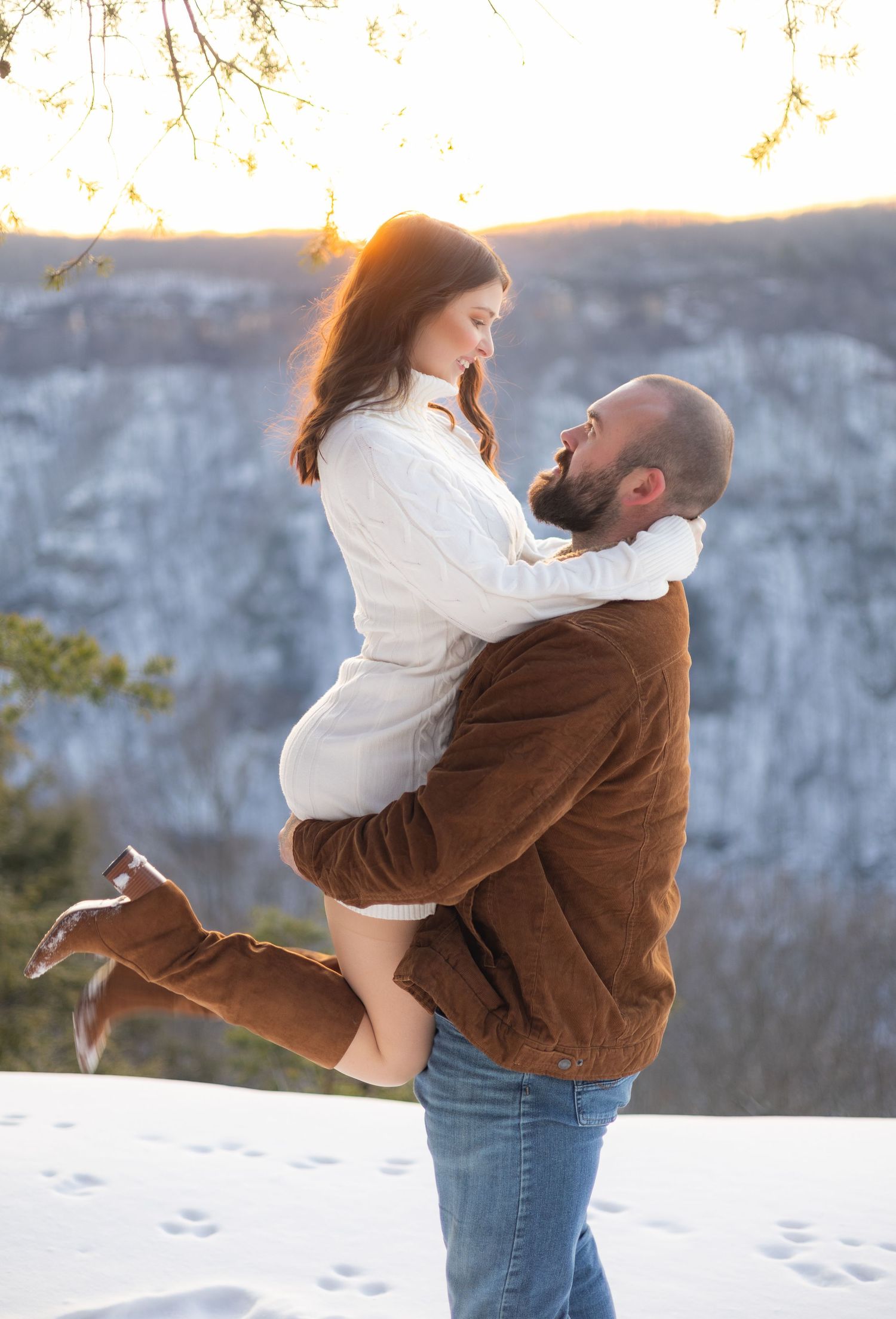 Bearded man lifting smiling woman in white sweater at snowy scenic overlook during golden sunset.