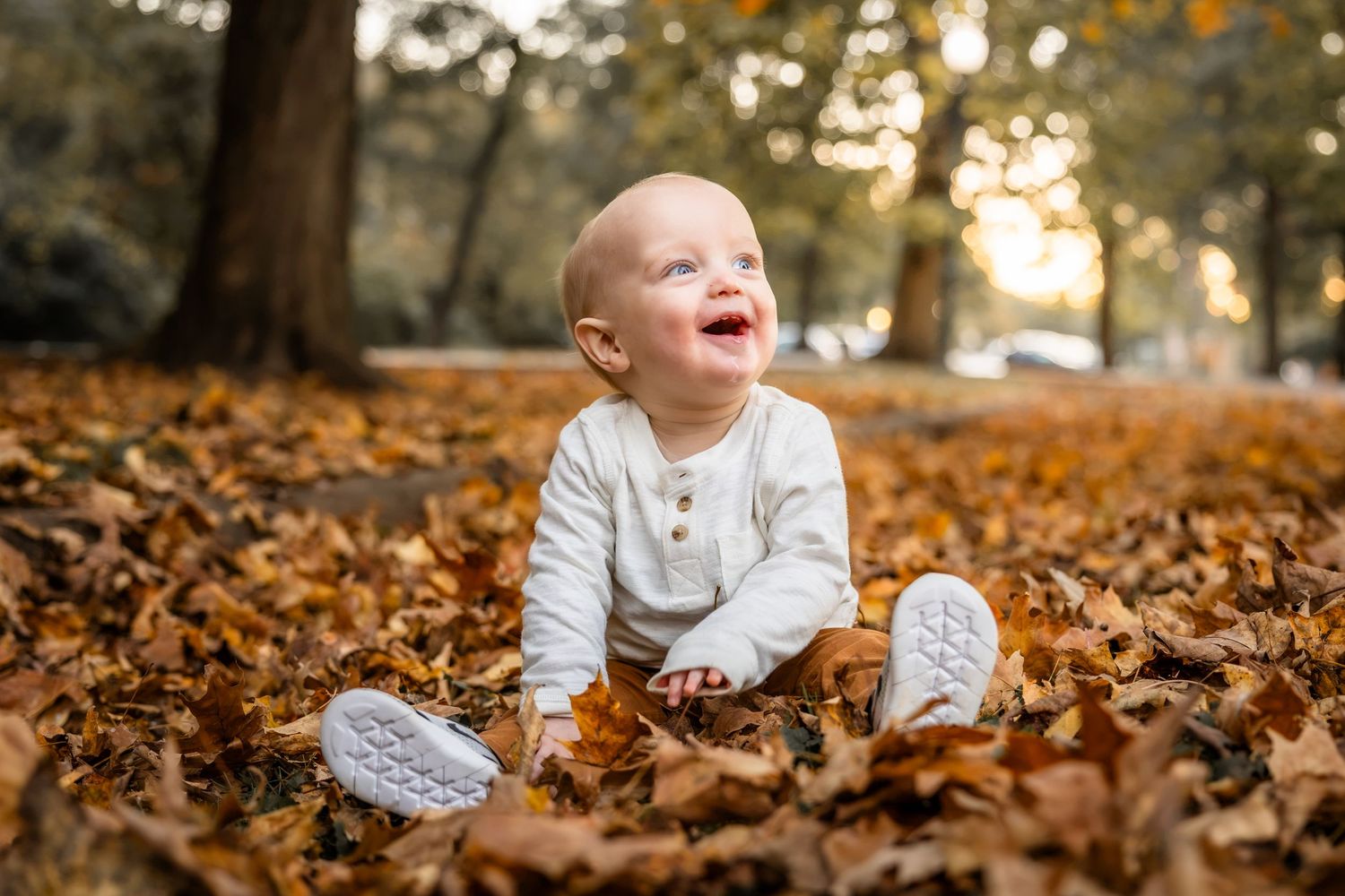 Smiling baby in a white outfit and white shoes sitting on a carpet of orange autumn leaves in a sunlit park.