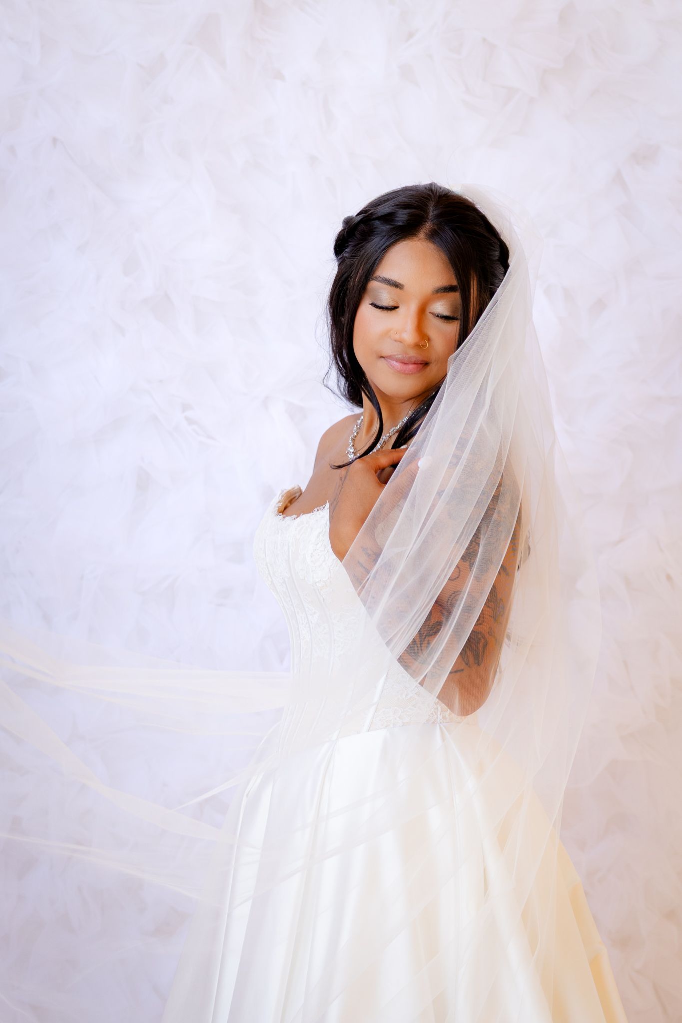 Elegant bride in white strapless gown with flowing veil poses with eyes closed gracefully.