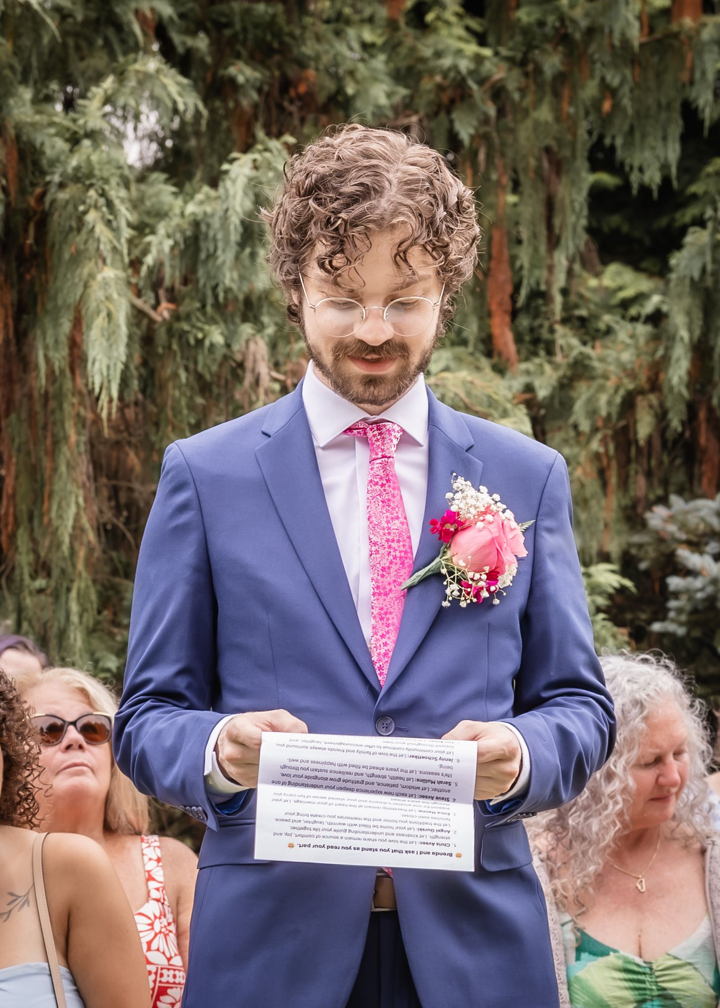 Man in blue suit with pink tie and boutonniere reads vows at outdoor wedding ceremony.