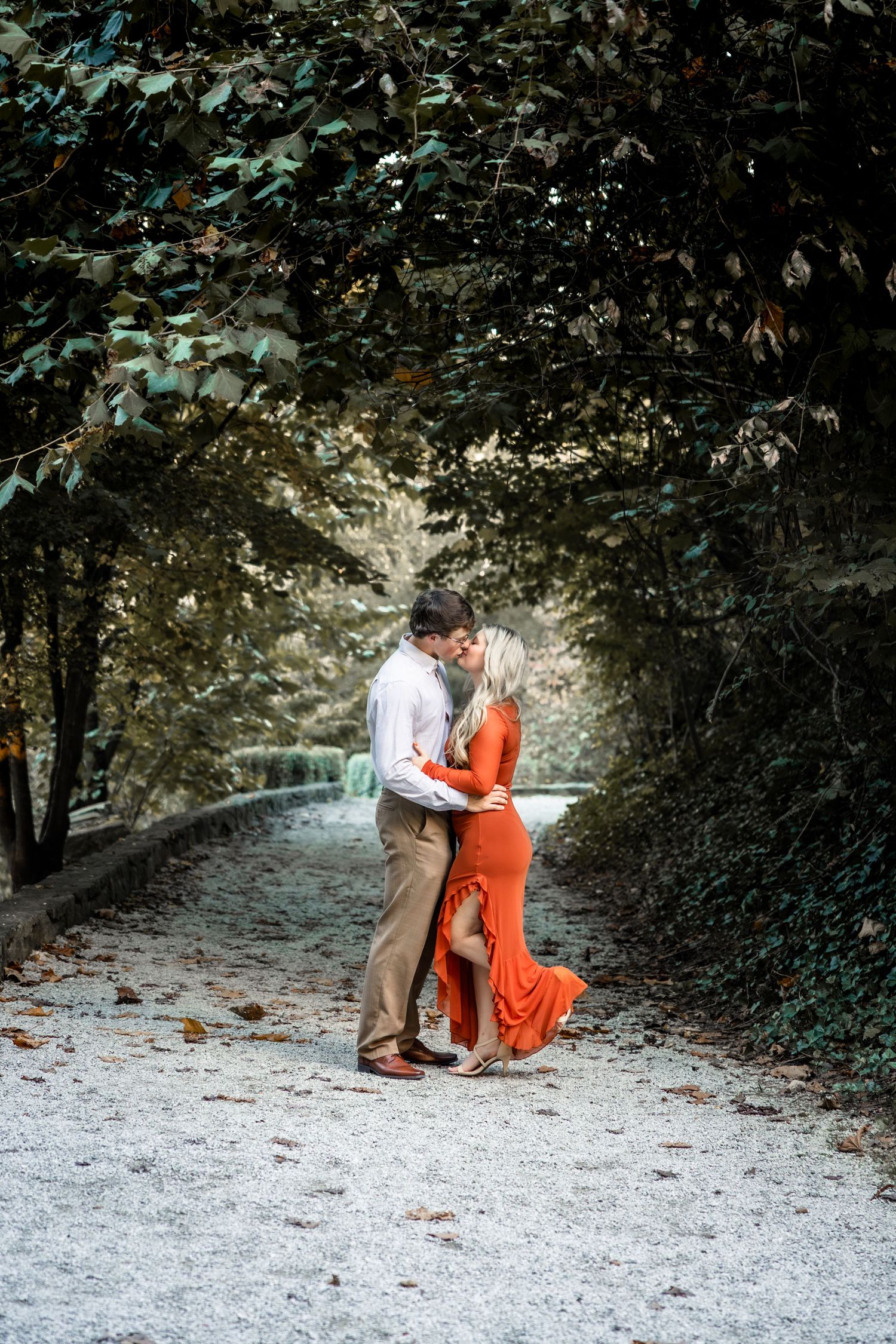 Couple sharing a romantic kiss on a tree-lined gravel path, woman in elegant orange gown, man in white shirt and tan pants.