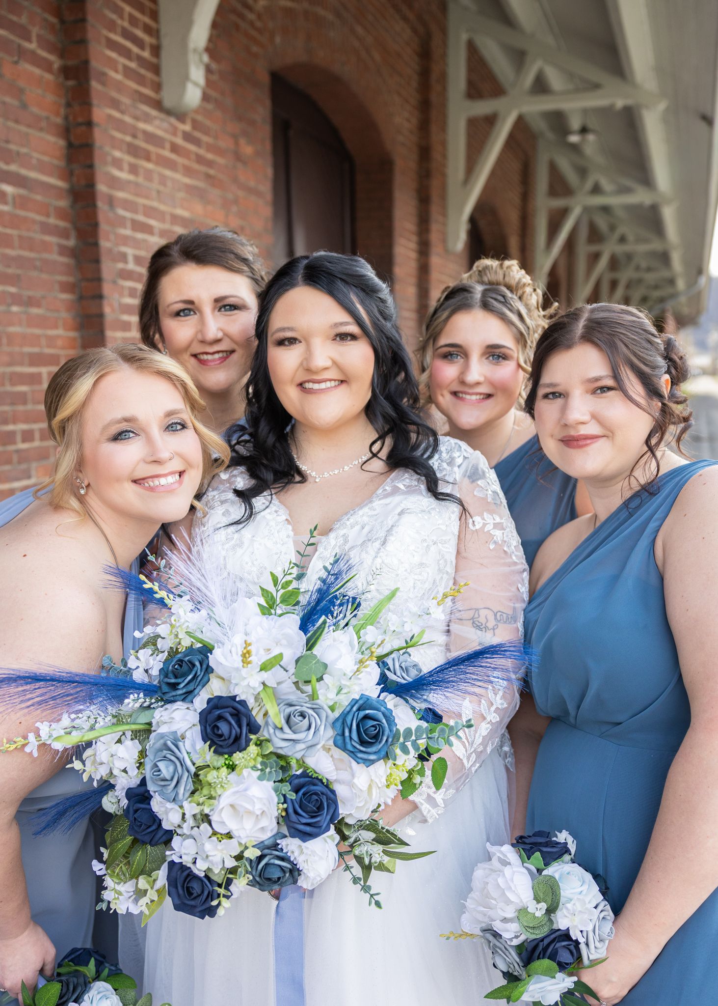 Bride in white lace gown holds blue rose bouquet surrounded by four bridesmaids in blue dresses outside brick building.