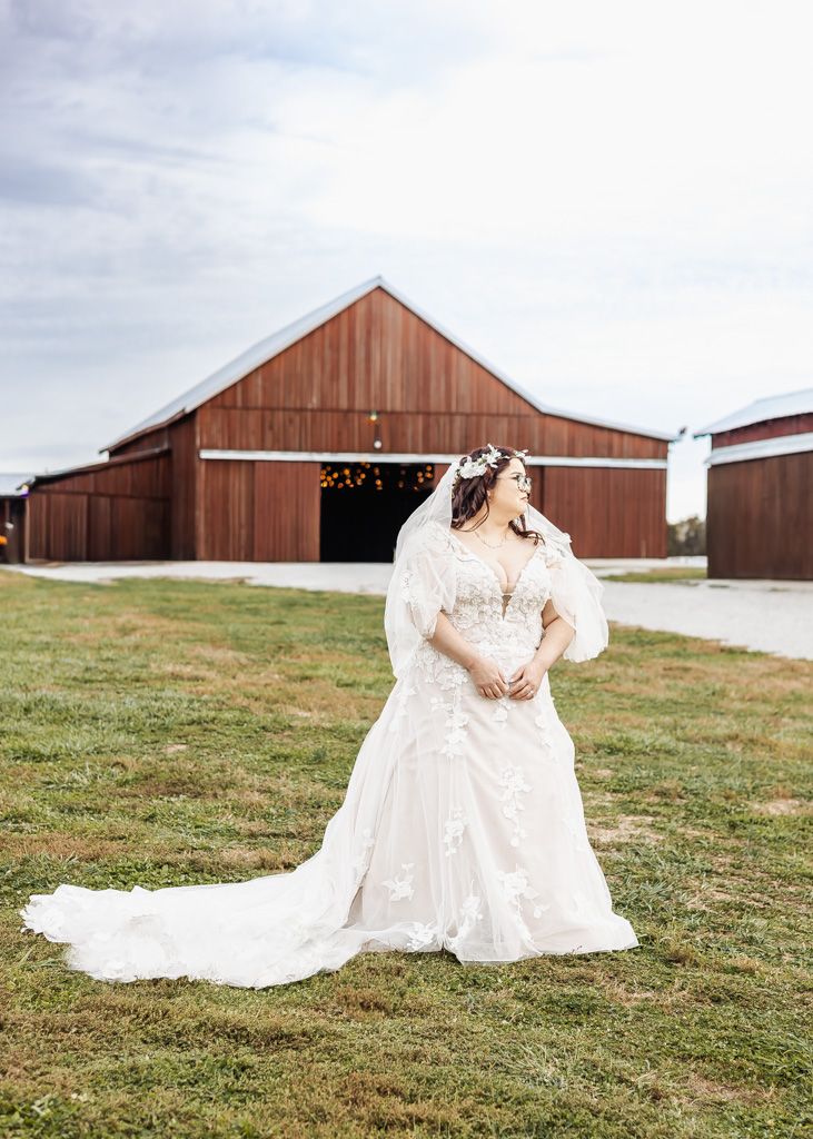 Bride in a white lace wedding gown with veil poses in front of a rustic red barn on a green lawn.
