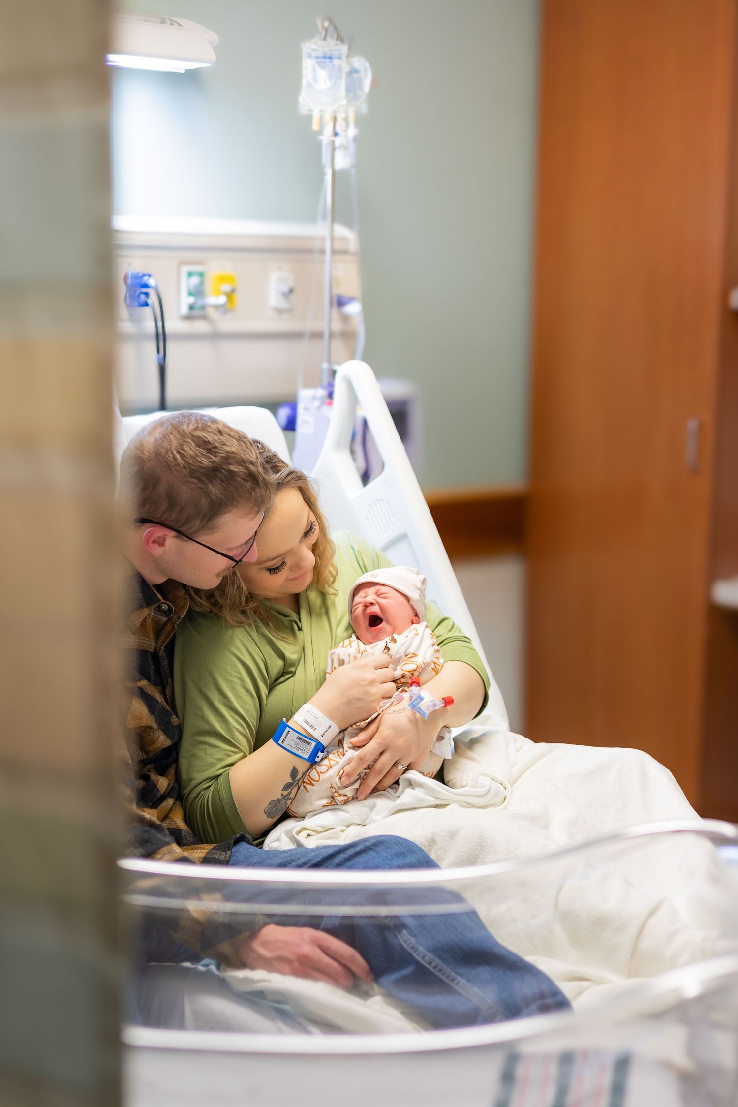 New parents in hospital room lovingly gazing at their crying newborn baby together.