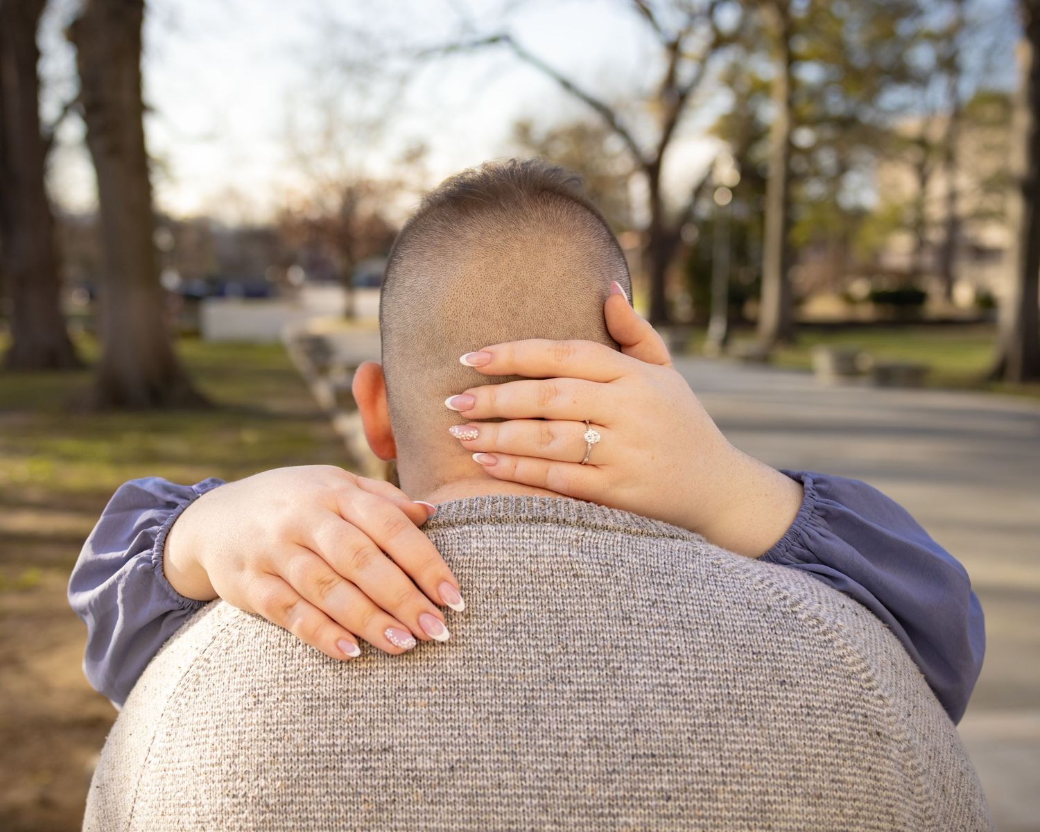 Close-up of woman's hand with engagement ring resting on man's shoulder outdoors.