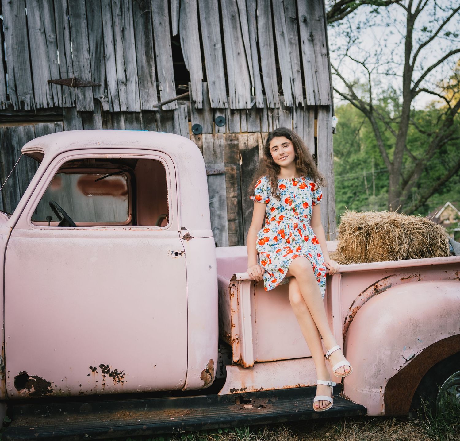 Young girl in floral dress sitting on rustic pink pickup truck bed near old barn with hay bale.