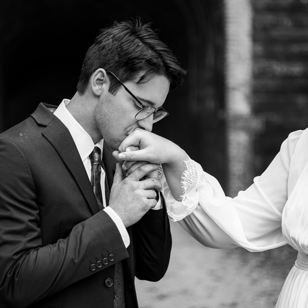 Groom in dark suit kissing bride's hand in romantic black and white wedding portrait.