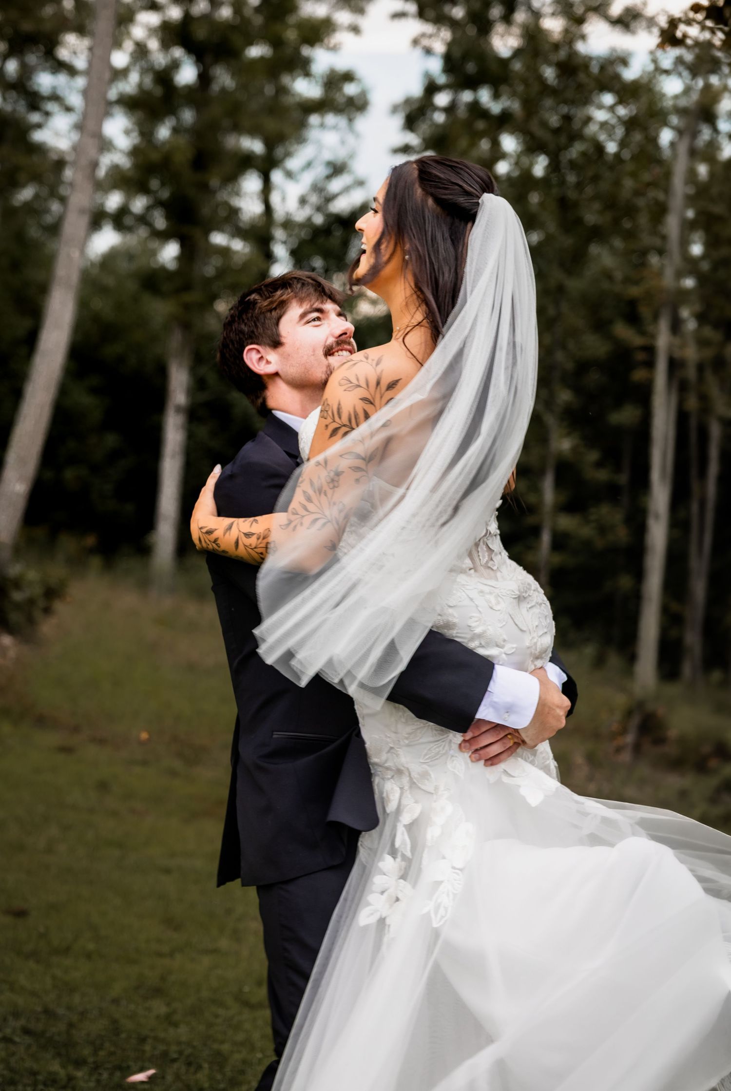 Groom in black tuxedo lifts bride in white lace gown and veil, both laughing joyfully outdoors among trees.
