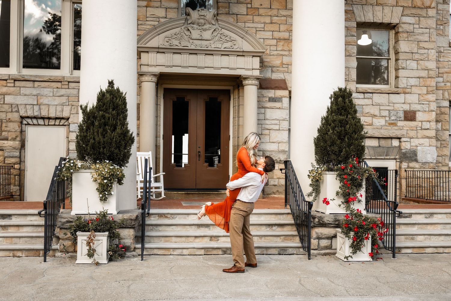 Couple embracing in front of elegant stone mansion entrance with evergreen topiaries and red floral accents.