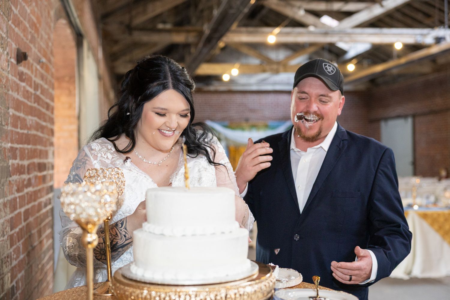 Bride and groom laughing together while cutting their white two-tier wedding cake at rustic indoor reception.