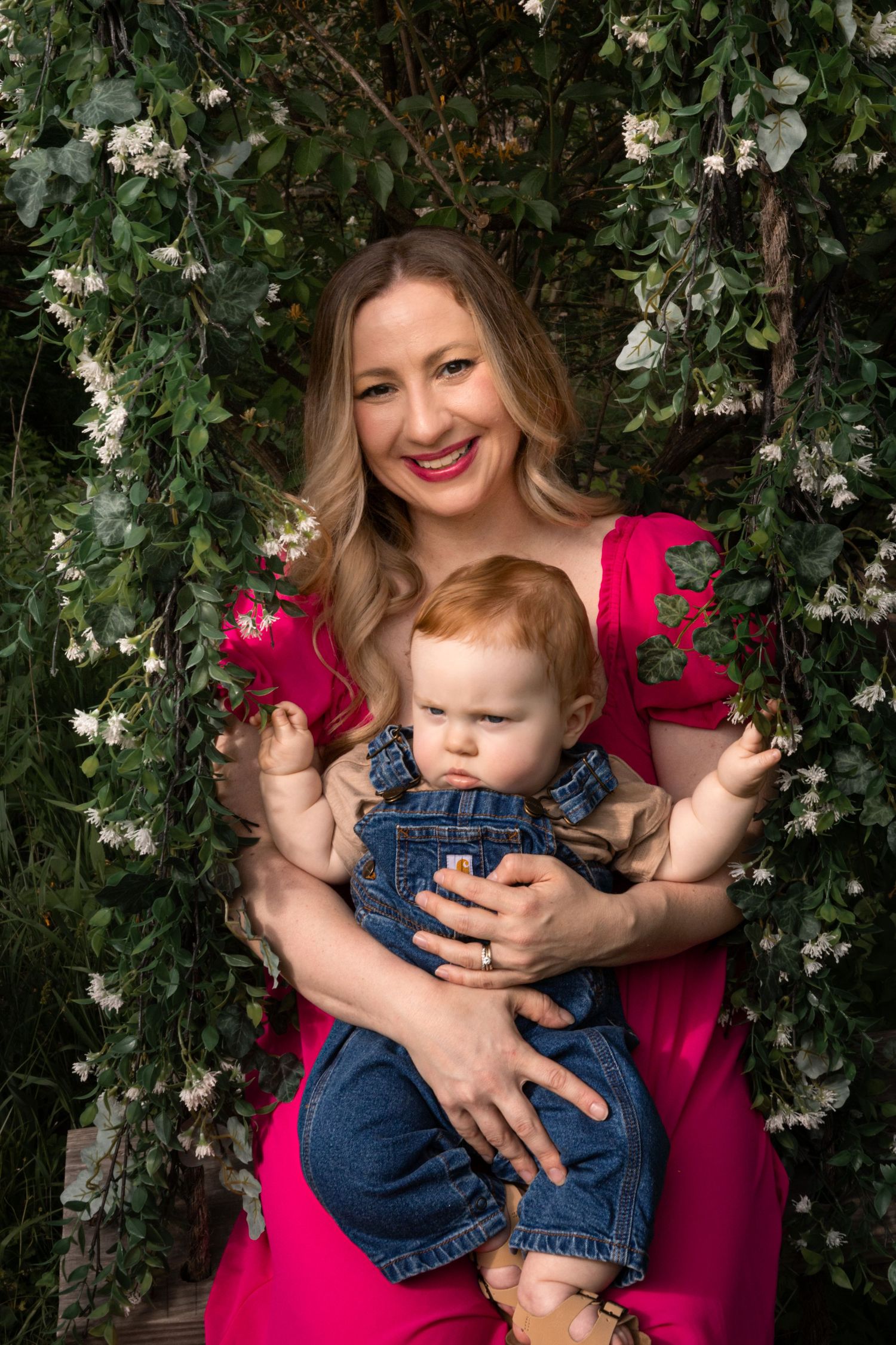 Smiling mother in pink dress holding red-haired baby on floral swing backdrop.