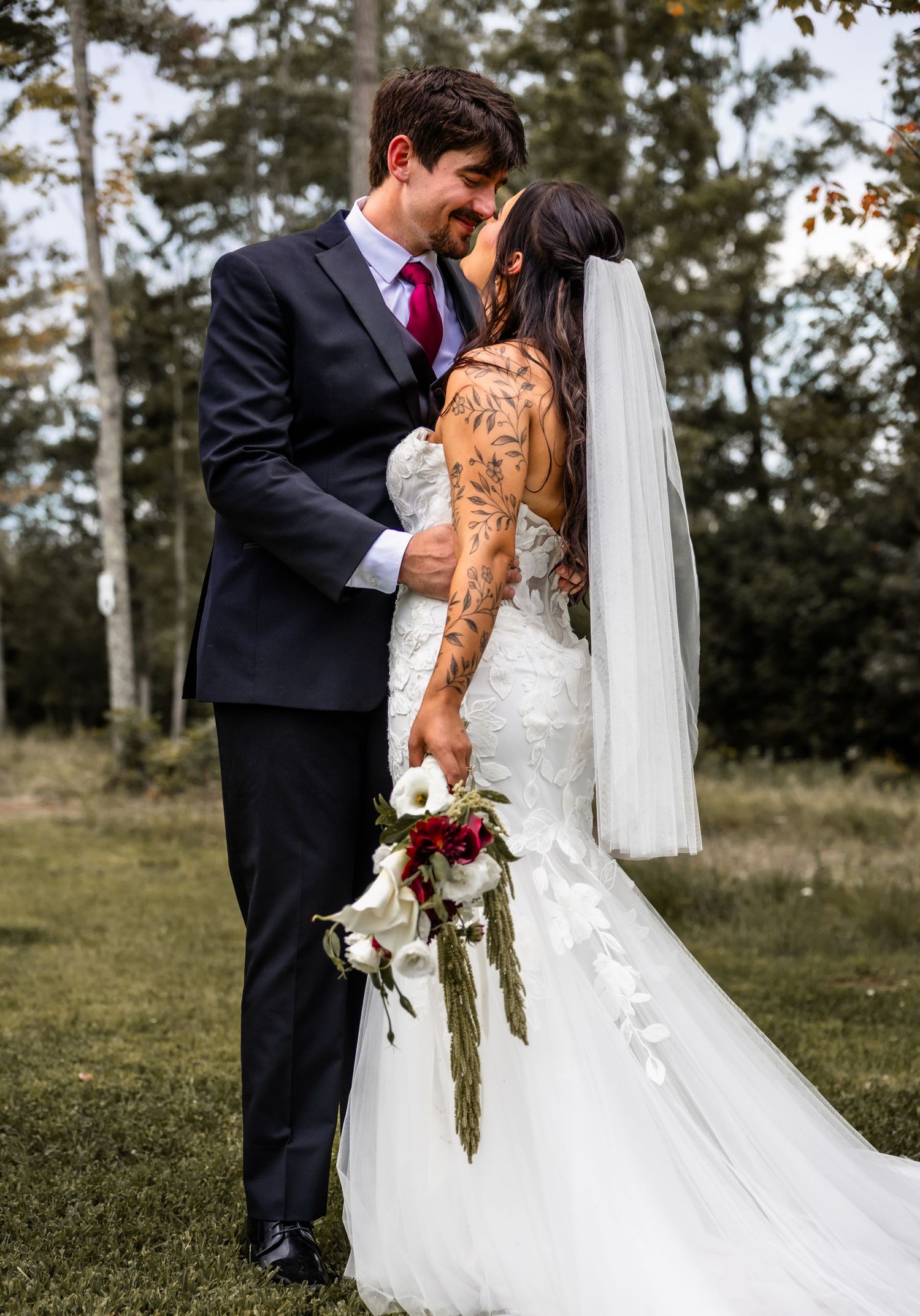 A bride and groom share a romantic moment outdoors, bride wearing lace gown with veil and holding red and white bouquet.
